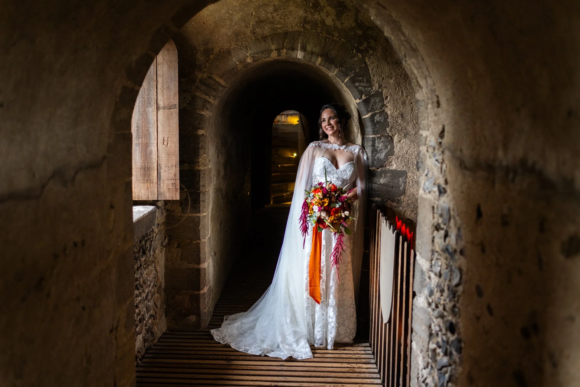 Bride in a white wedding dress holding a colorful bouquet standing in a stone arched hallway.