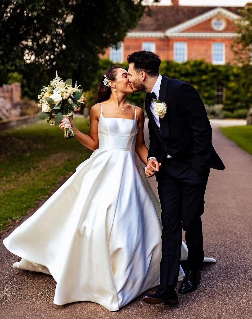 A bride and groom share a kiss while holding hands on a wedding day, with the bride holding a bouquet and wearing a white wedding gown, and the groom in a black tuxedo with a white rose boutonniere, outdoors near a brick building.