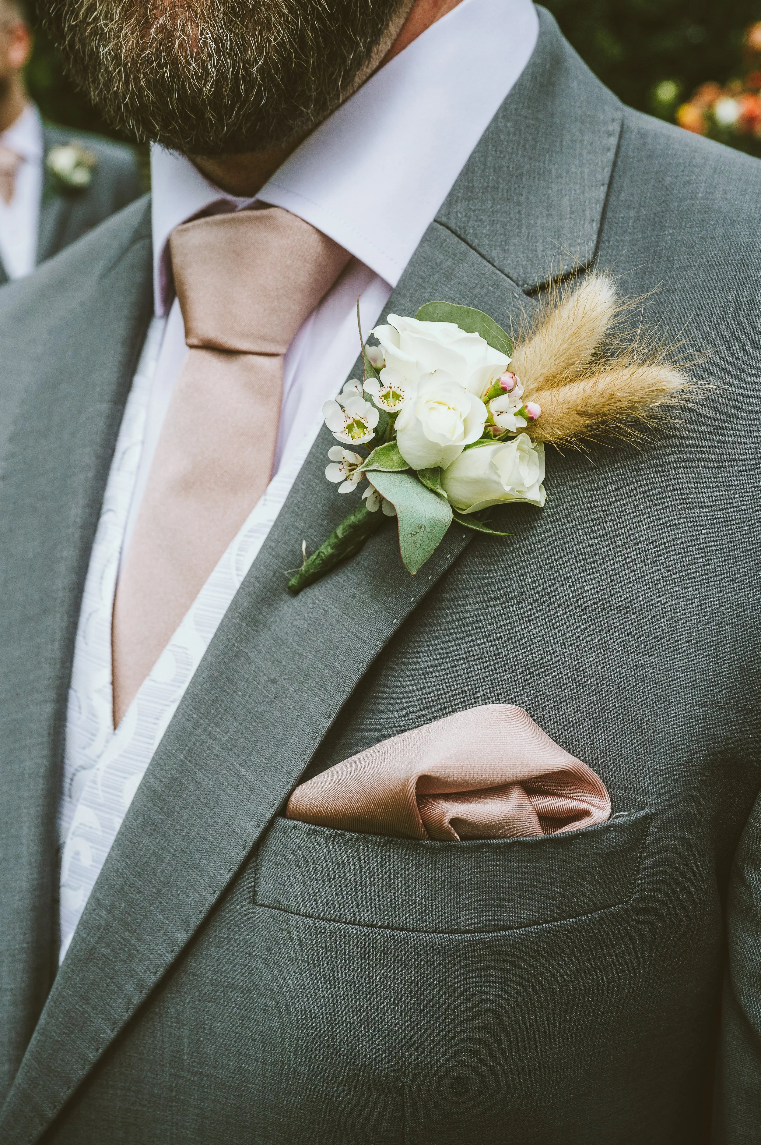 Close-up of a man's suit with a boutonnière that includes white flowers, greenery, and wheat, with a peach-colored tie and pocket square.