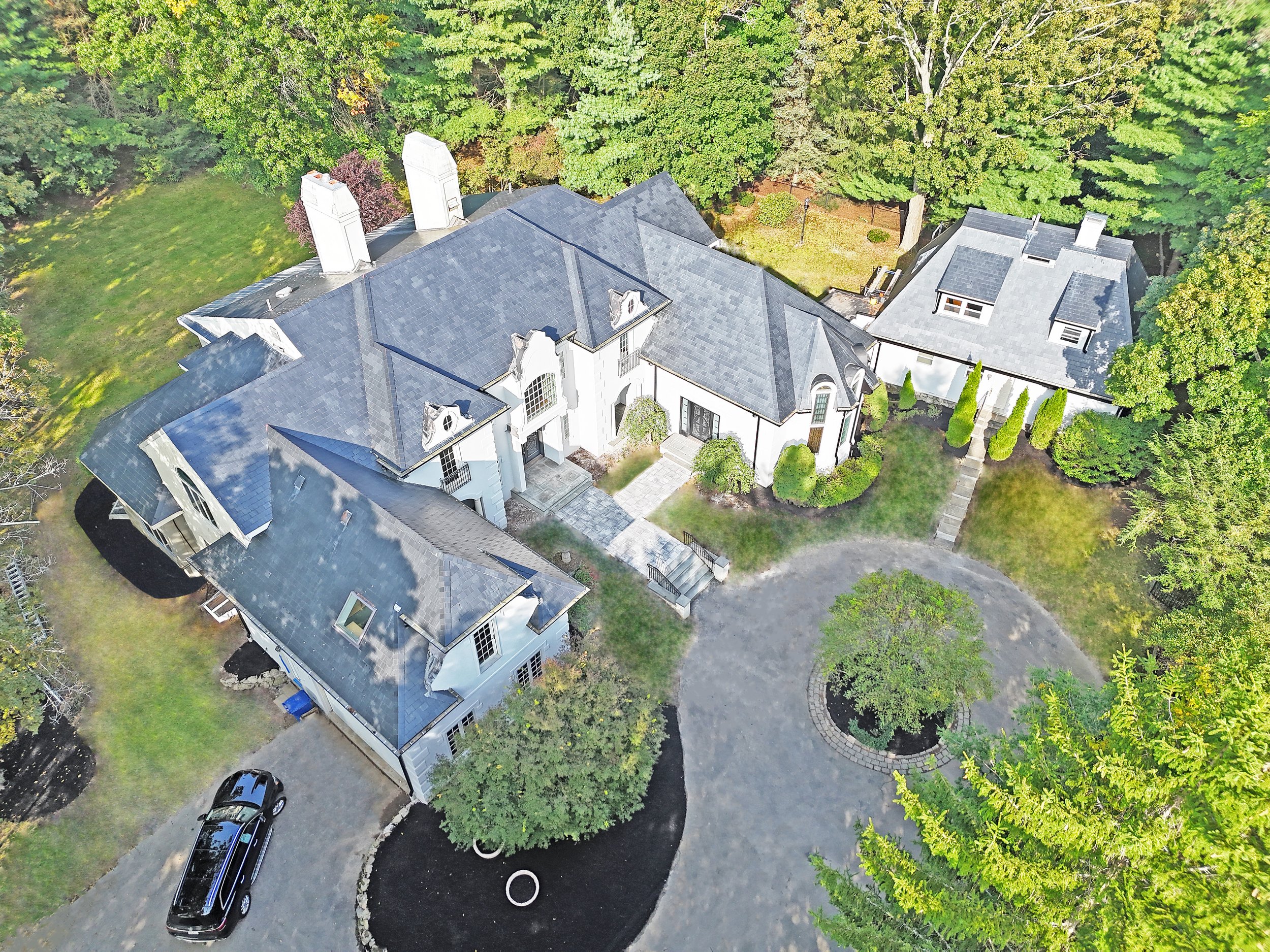 An aerial view of a large, elegant house with a dark gray roof, surrounded by trees and a landscaped yard with a circular driveway and a small garden area.