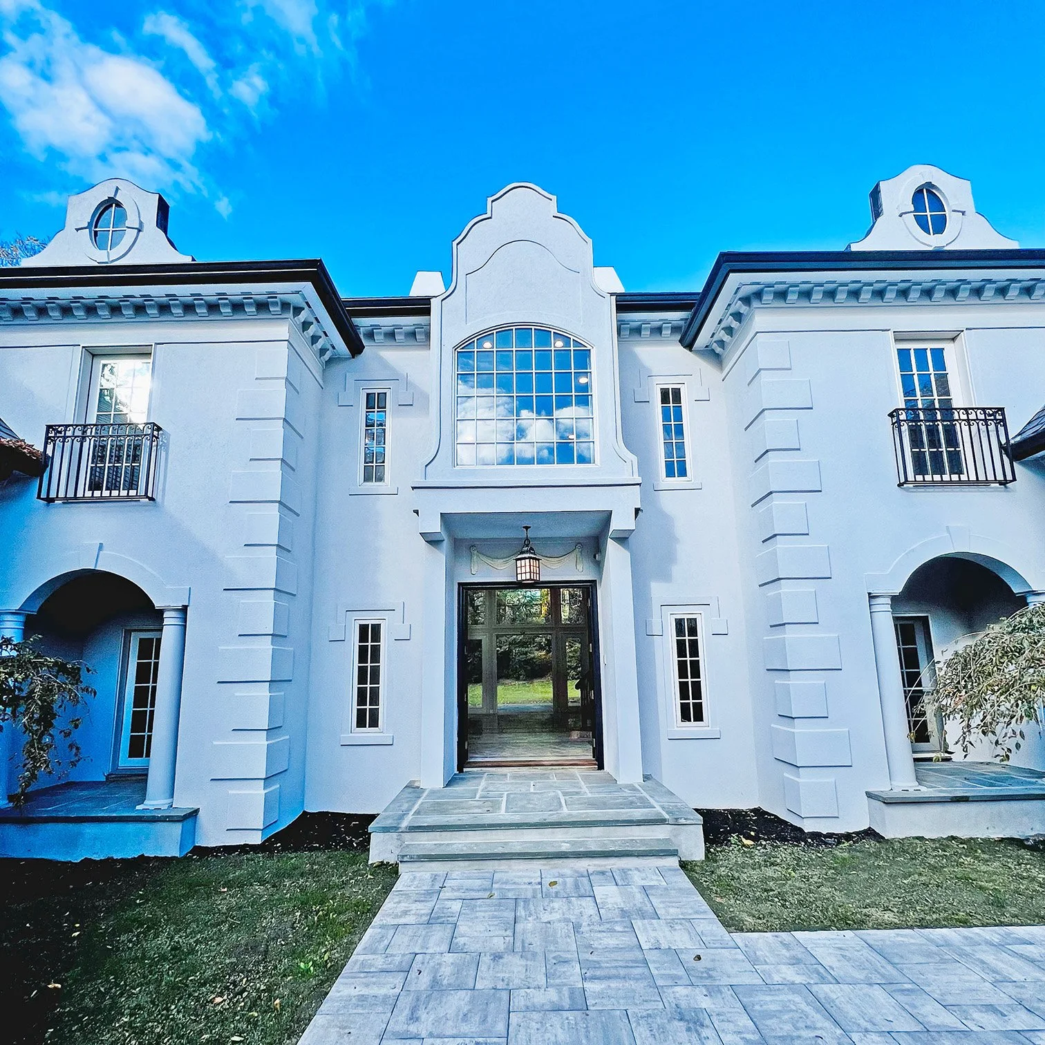 Exterior view of a white house with a large arched window, small balconies, and a pathway leading to the entrance under a blue sky with clouds