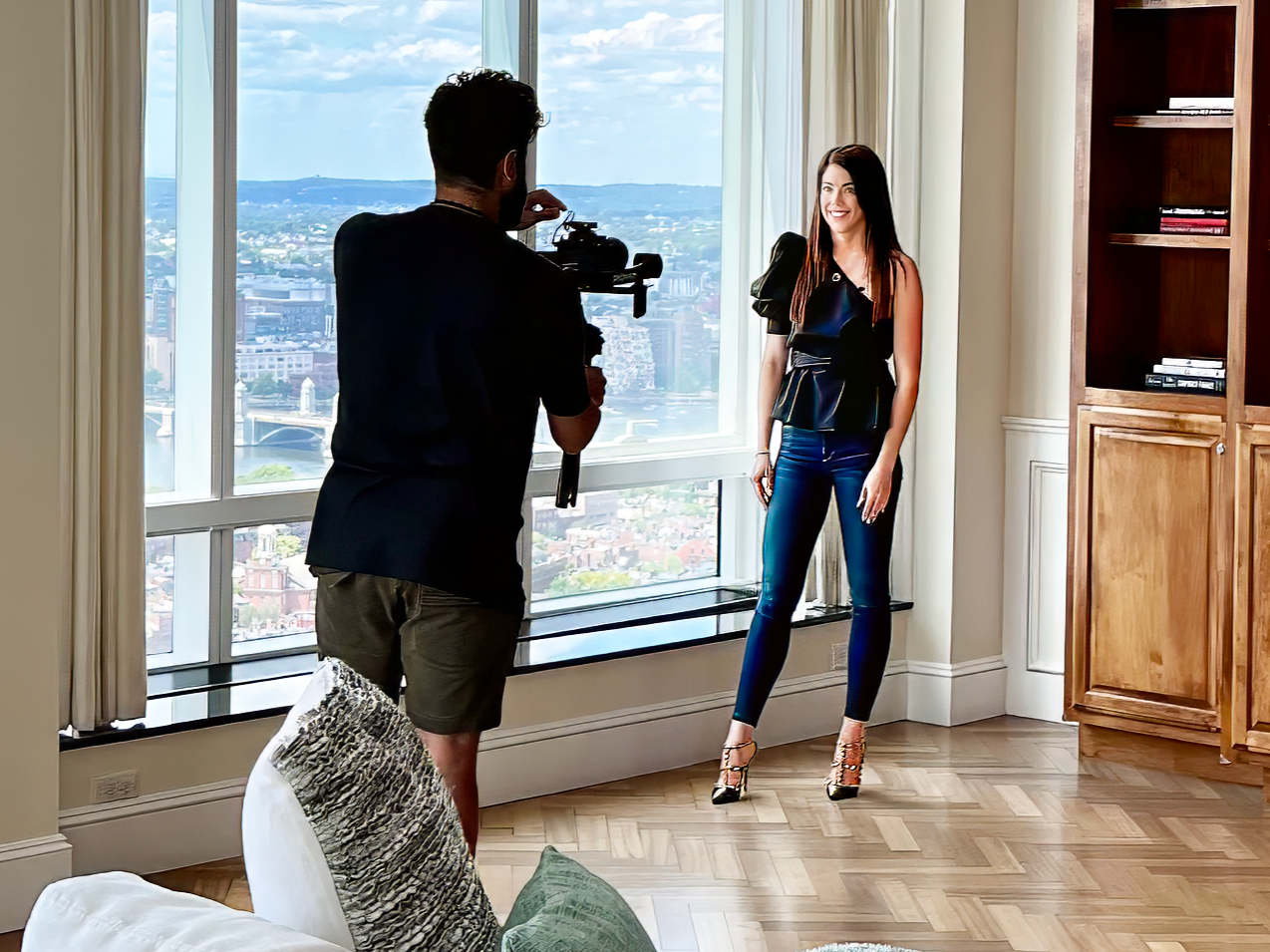 A woman is being filmed by a man with a camera in a room with large windows and a city view.