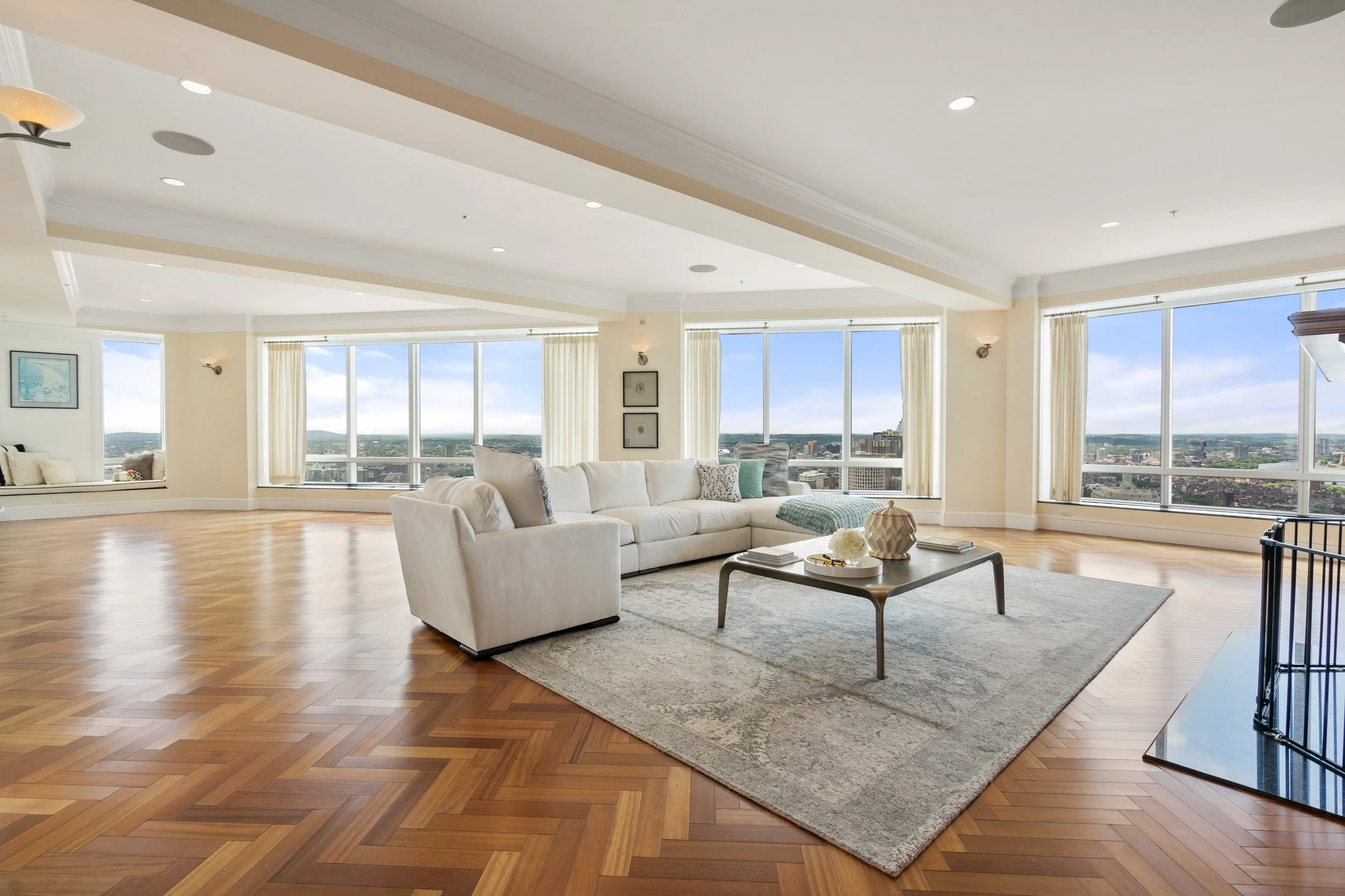 Spacious living room with large windows showing city view, white sectional sofa, coffee table, and hardwood floors.