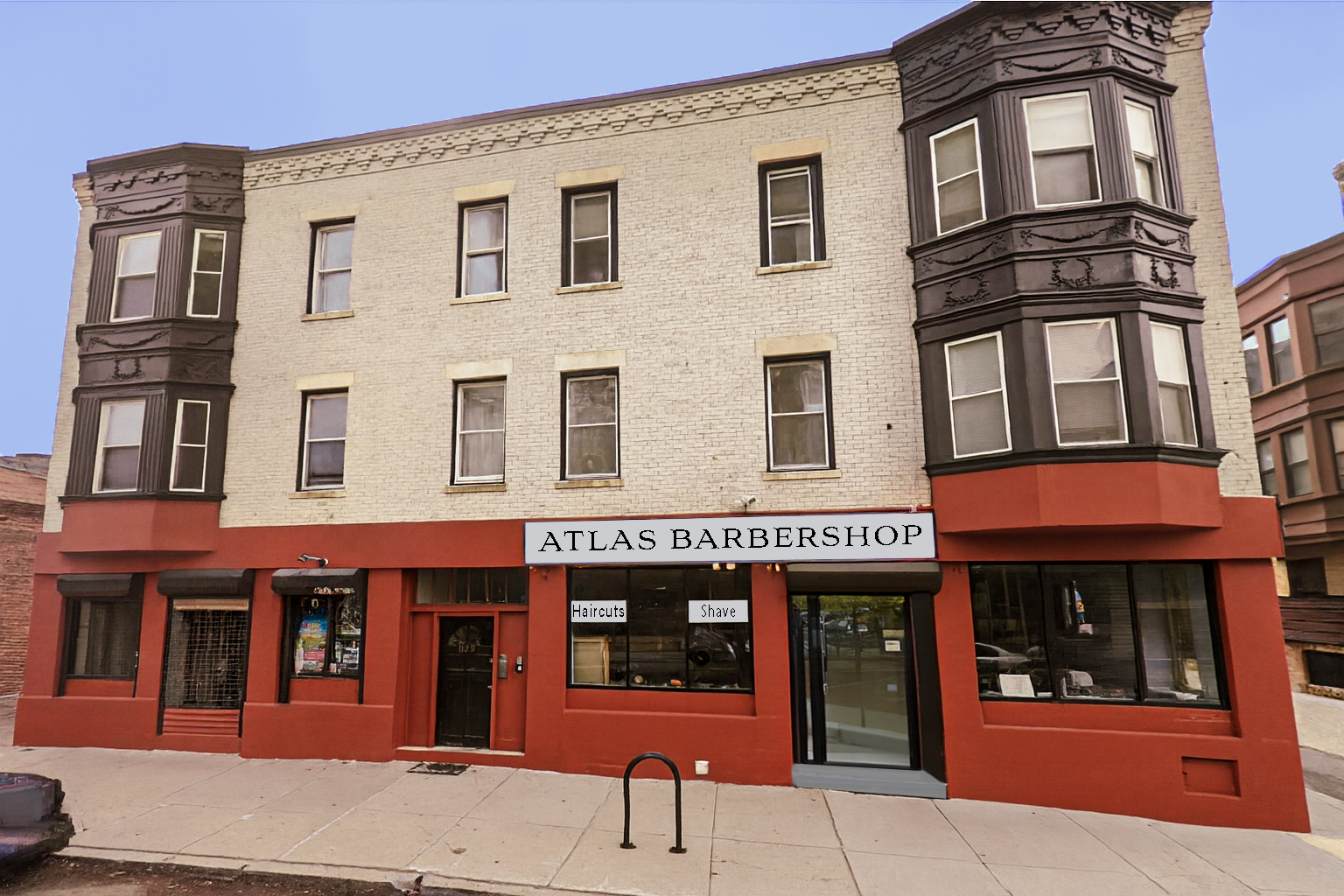 Three-story building with a barbershop on the ground floor, with beige brick exterior, black bay windows on the upper floors, and a red facade on the bottom. The barbershop sign reads "ATLAS BARBERSHOP" and has smaller signs for "Haircuts" and "Shave" in the windows.