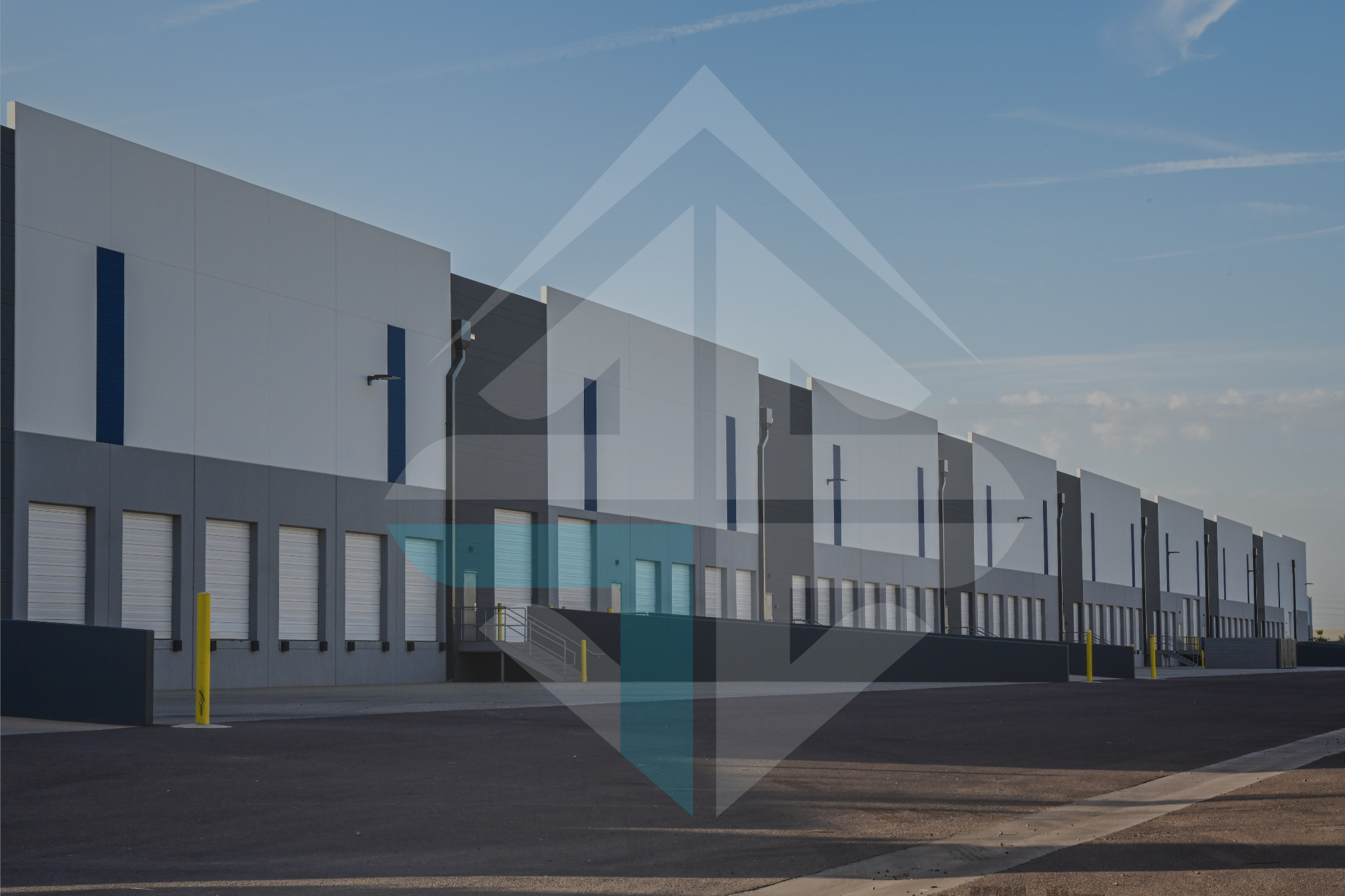 Exterior view of a large industrial warehouse building with multiple loading docks, yellow safety bollards, and an empty parking area under a clear blue sky.