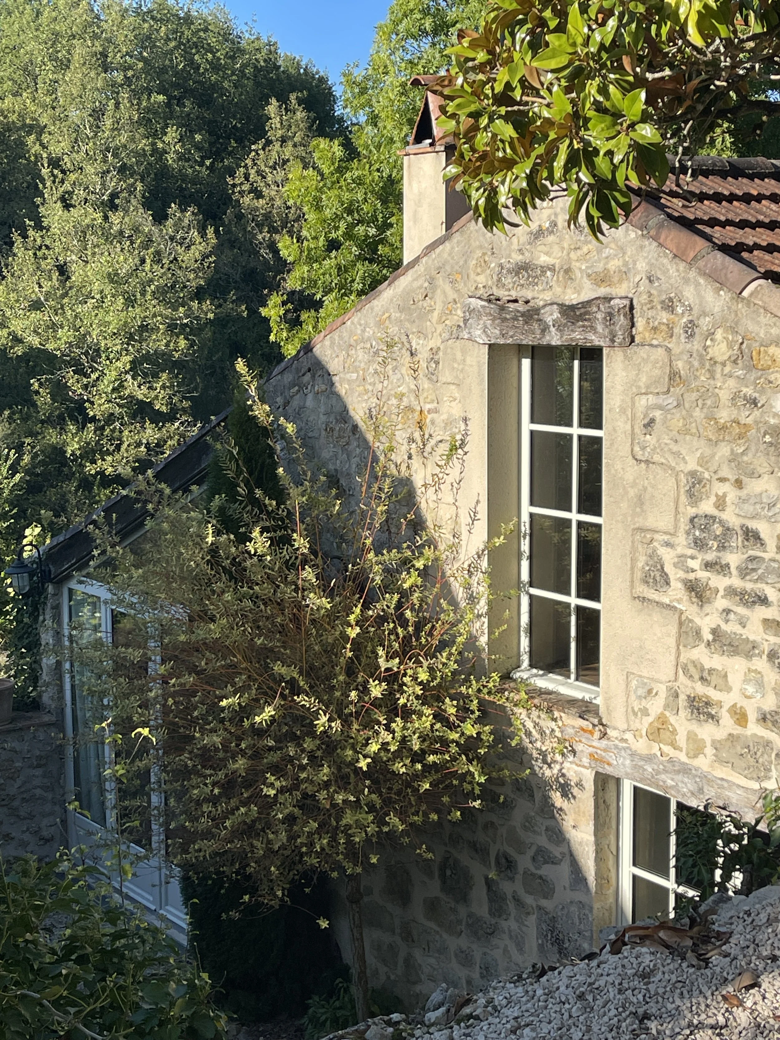 A stone cottage with a pitched roof and large white-framed windows, surrounded by green trees and bushes under a clear blue sky.
