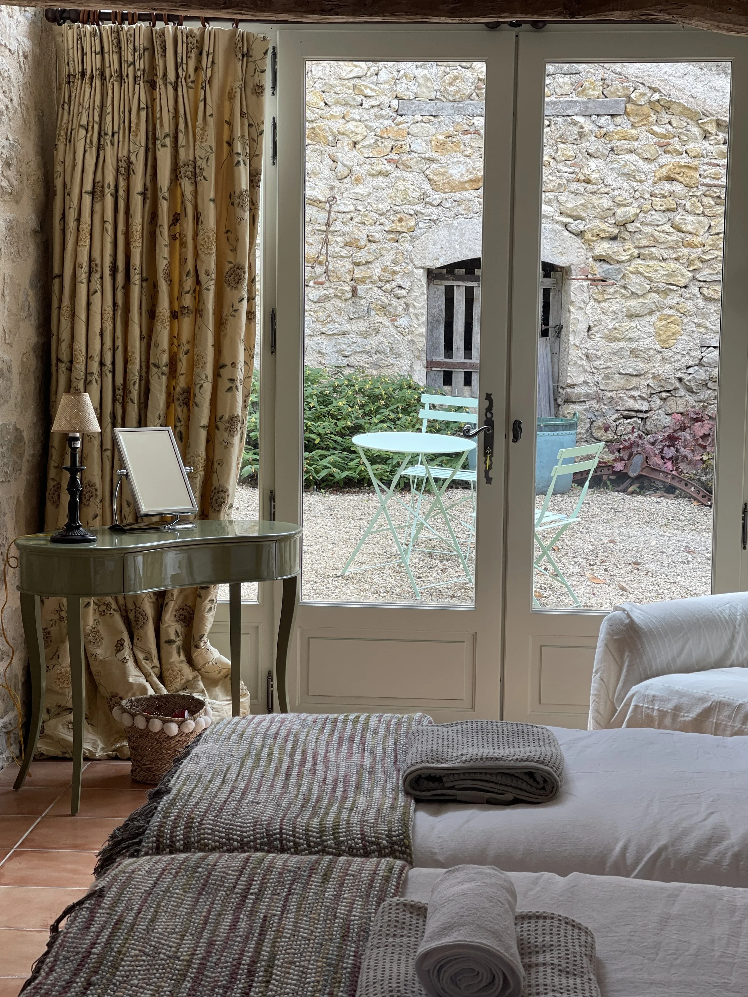 Indoor bedroom with a view of a small outdoor patio. The room has a French door with floral curtains, a small green table with matching chairs outside, and a bed with folded towels on top.