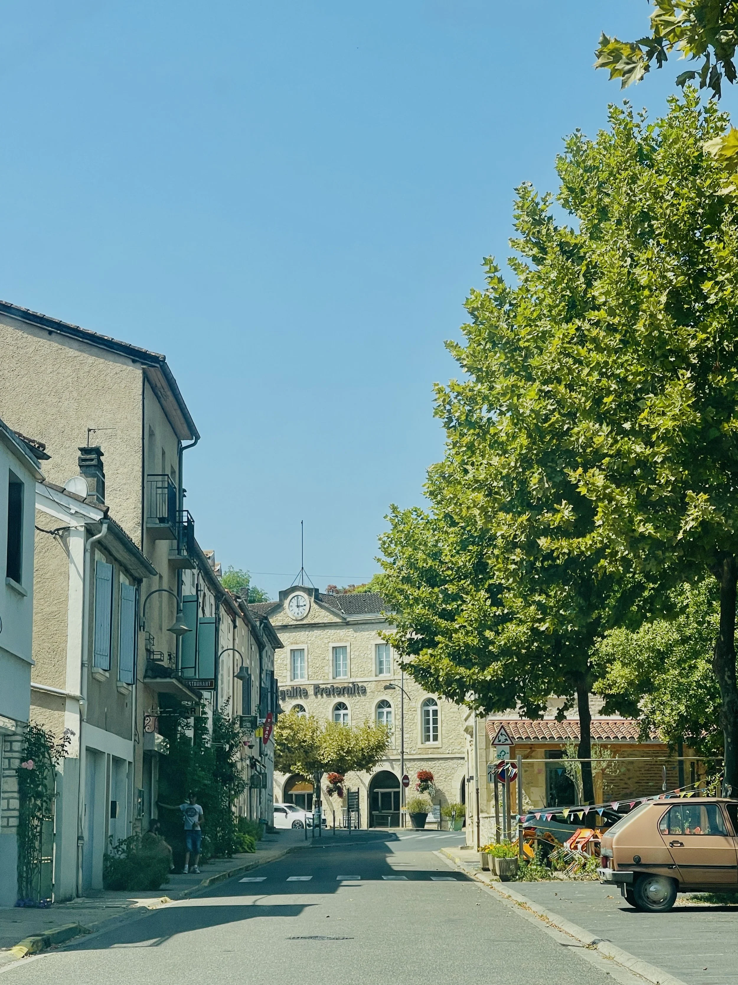 A quiet street in a small town with old buildings, green trees, and a clear blue sky, featuring a clock on a building and parked cars.
