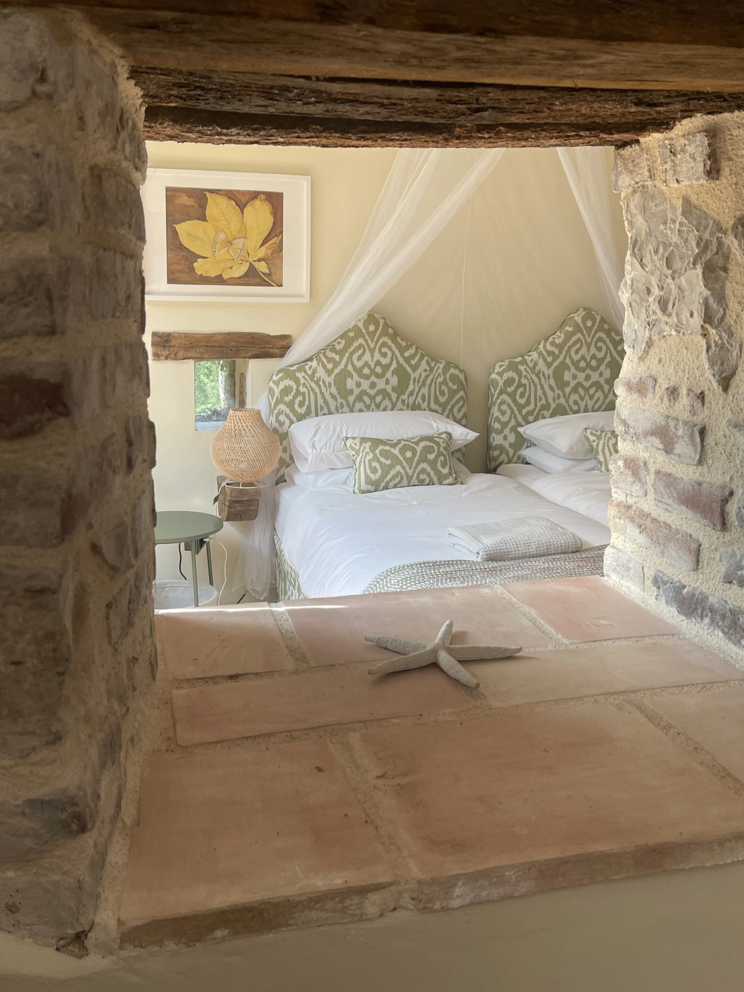 A cozy bedroom with a white bed featuring green patterned headboards and pillows, surrounded by rustic brick and wood accents, and decorated with a starfish on the windowsill.