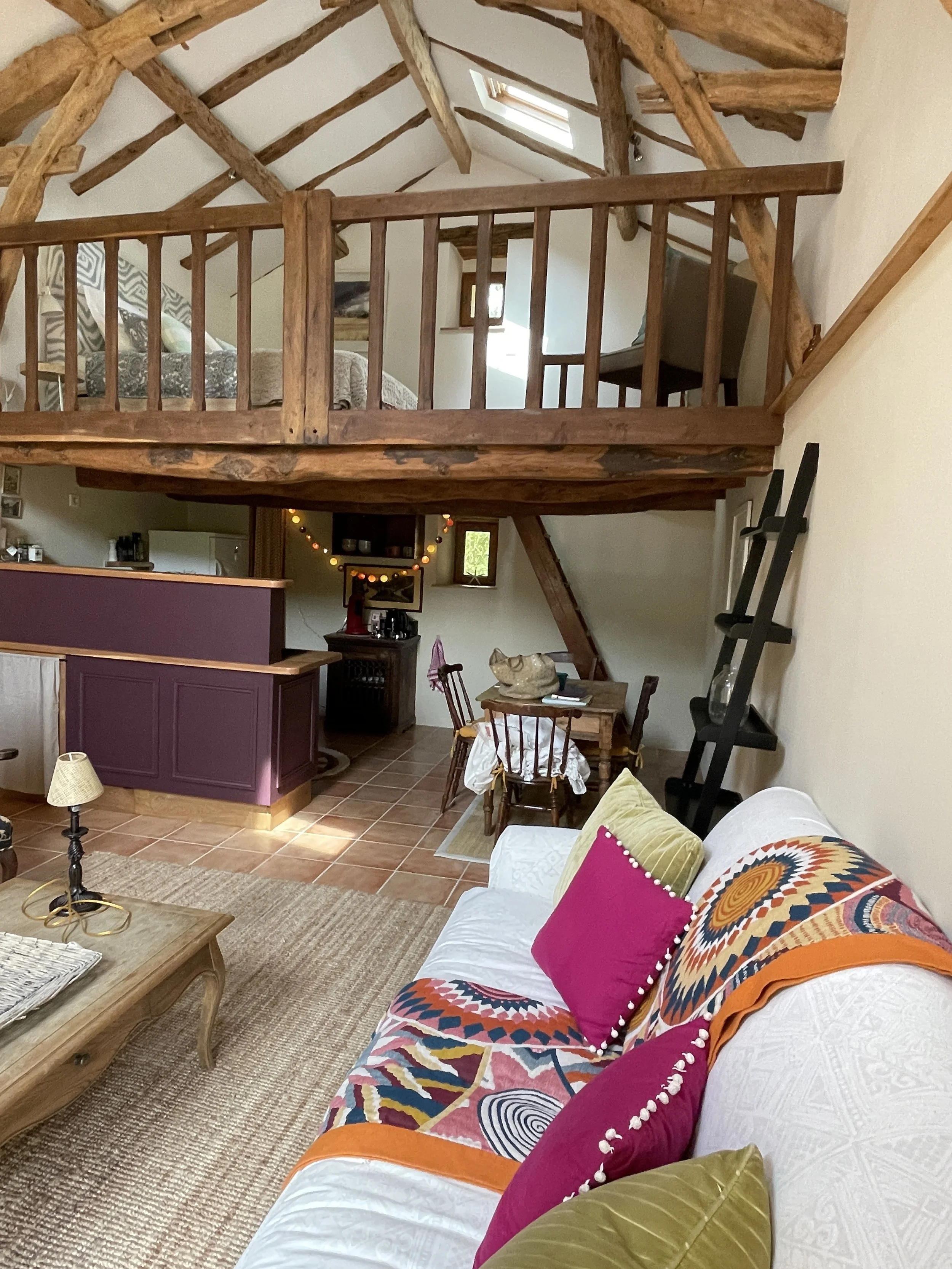 A cozy living room with a white sofa decorated with colorful pillows and a blanket, a wooden coffee table, and a tiled floor. There's a loft area with wooden railing, a bed, and a chair, accessible via angled staircase. The space has rustic wooden be