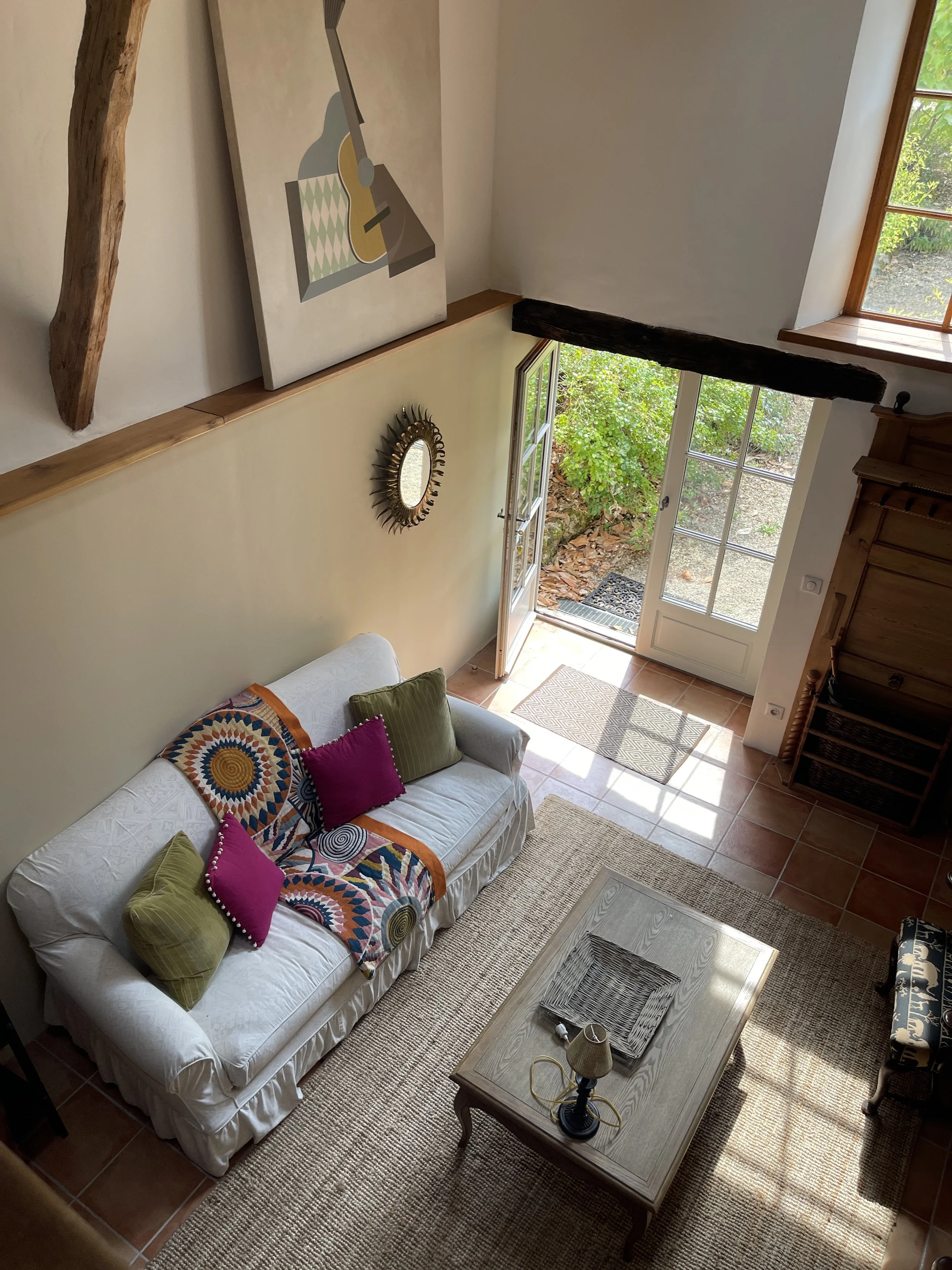 Living room with a white sofa decorated with colorful pillows and a patterned throw, a wooden coffee table with a lamp and decorative tray, a stone and tile floor, and large glass double doors opening to an outdoor area with greenery.
