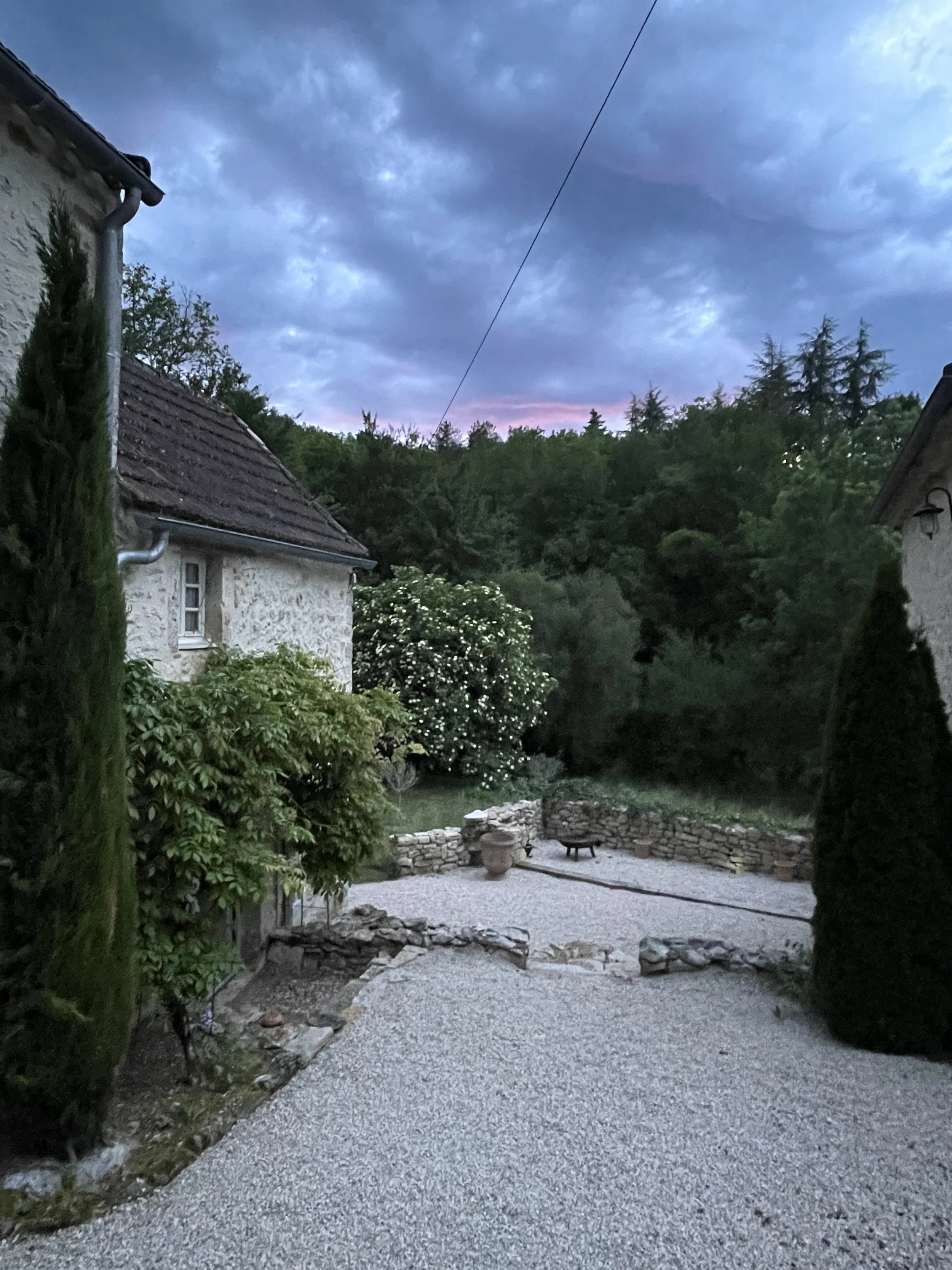 A gravel courtyard surrounded by stone walls, with a potted plant and a small fire pit, flanked by two stone houses with tiled roofs, green trees and bushes, and a cloudy sky at dusk.