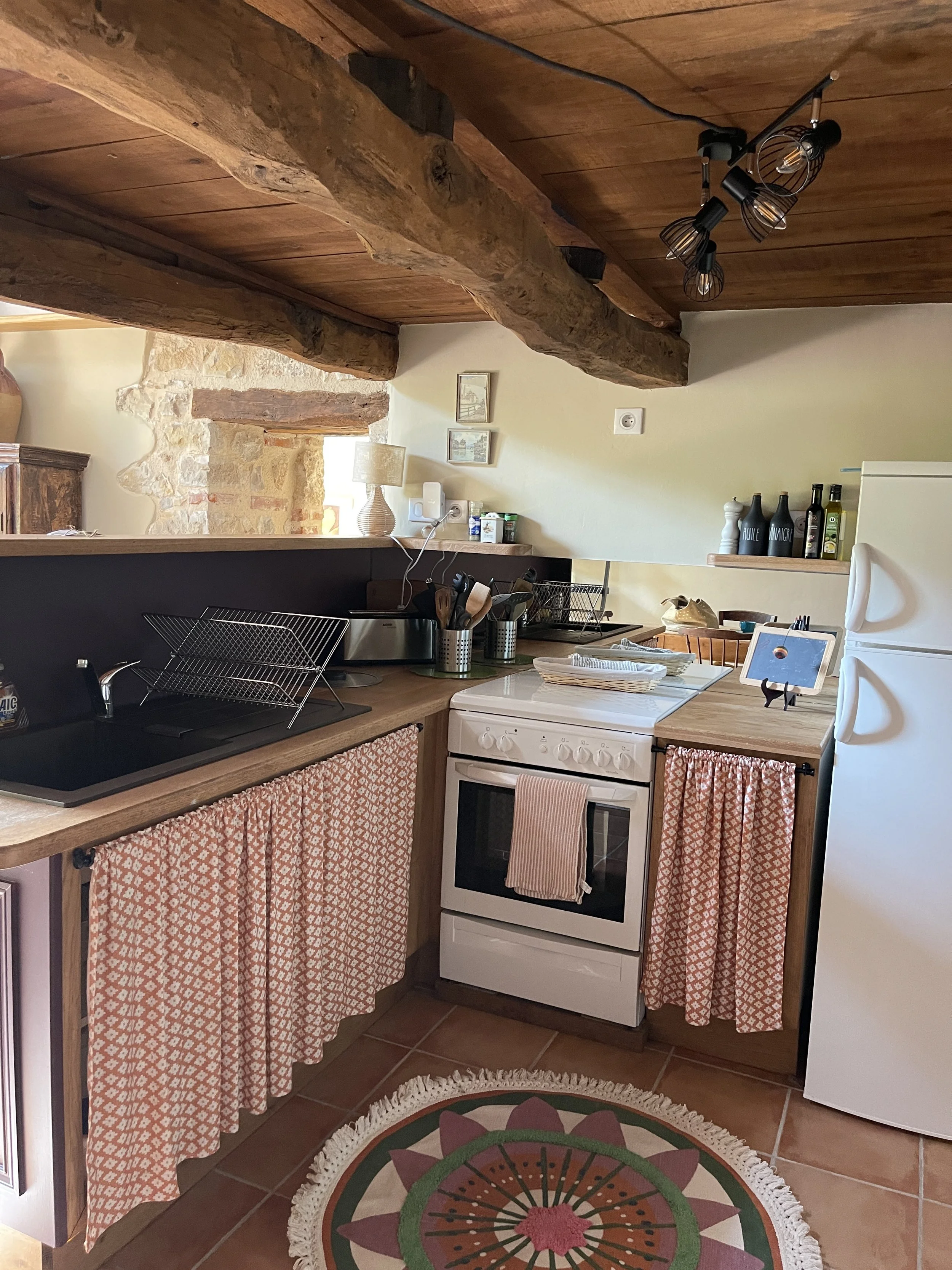 Cozy kitchen with wooden beams on the ceiling, a white stove with a pink towel hanging, a white refrigerator, and pink patterned curtains below the wooden countertops. There is a colorful round rug on the tile floor, and various kitchen items and dec