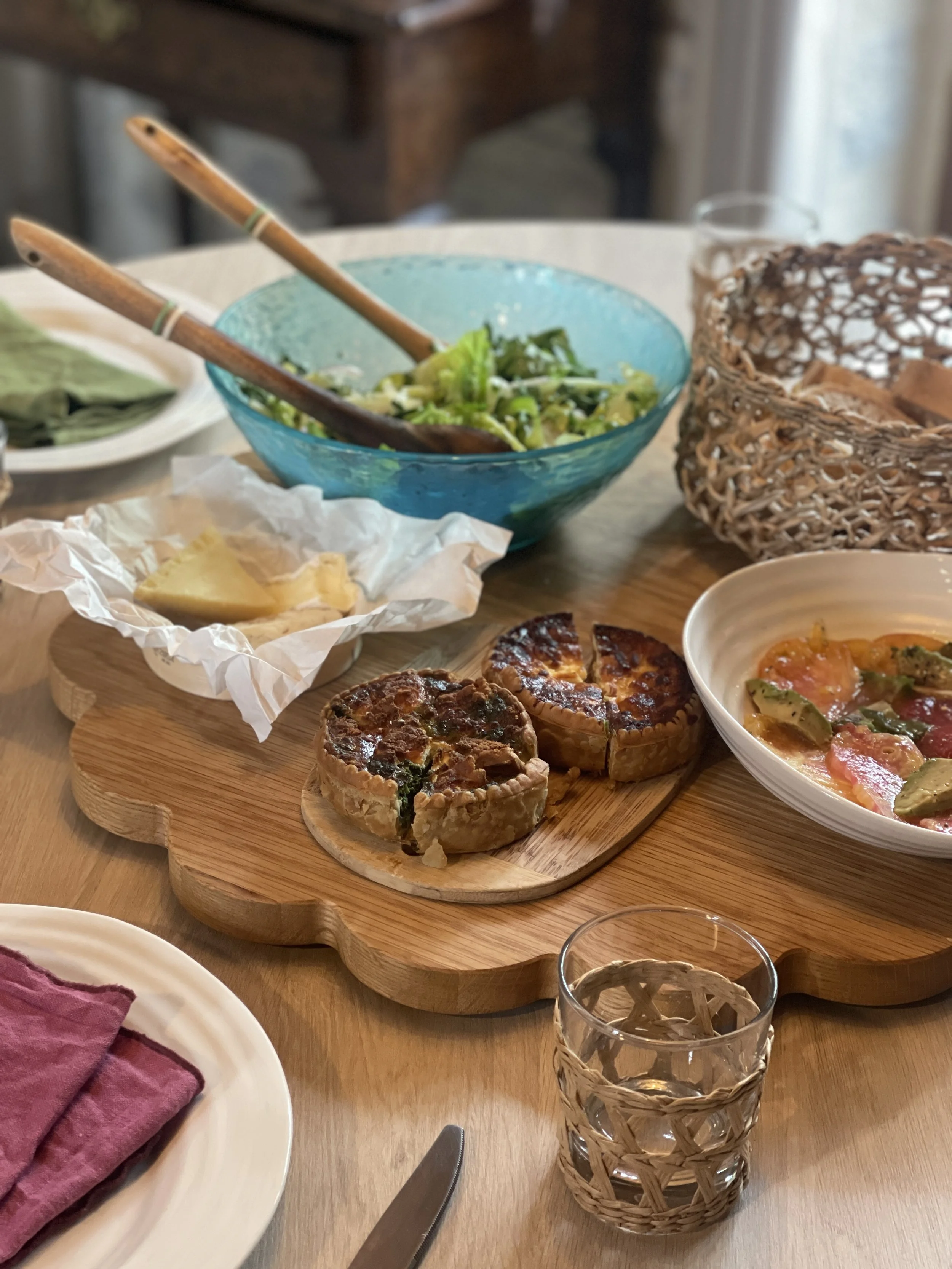 A table set with various dishes, including a bowl of salad, a cheese plate, two small quiches on a wooden serving board, a bowl of baked tomatoes and vegetables, a glass of water, and a woven basket for bread.
