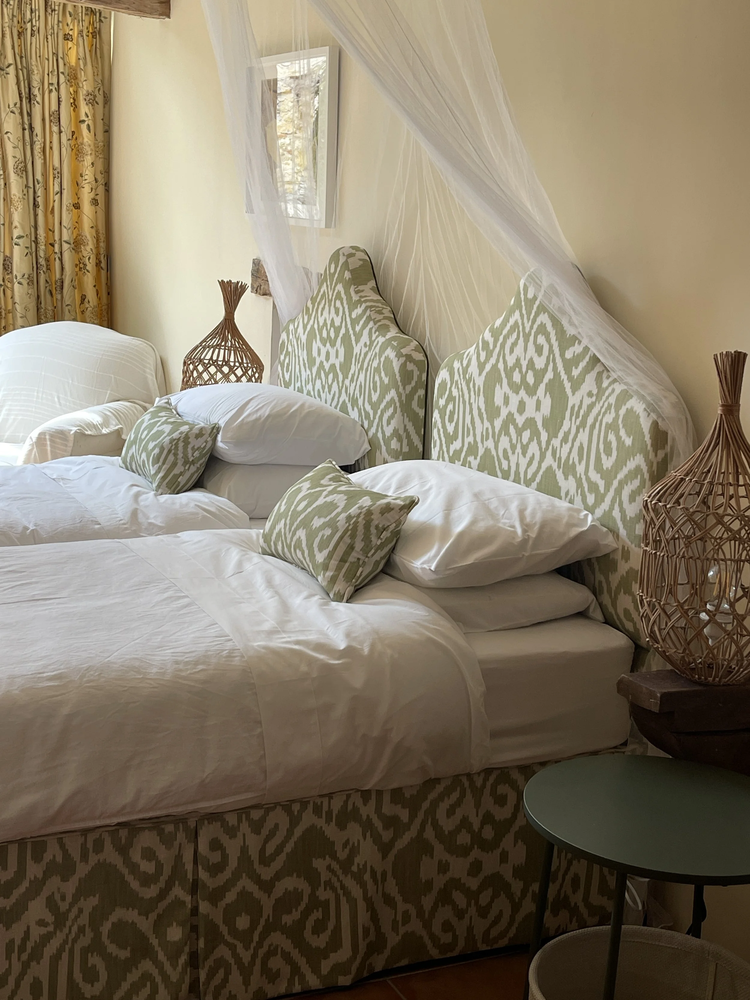 A bedroom with a large bed with white bedding, green and white patterned pillows and headboards, a mosquito net canopy, a wicker nightstand with a decorative wicker lamp, a window with yellow floral curtains, and a small green side table.