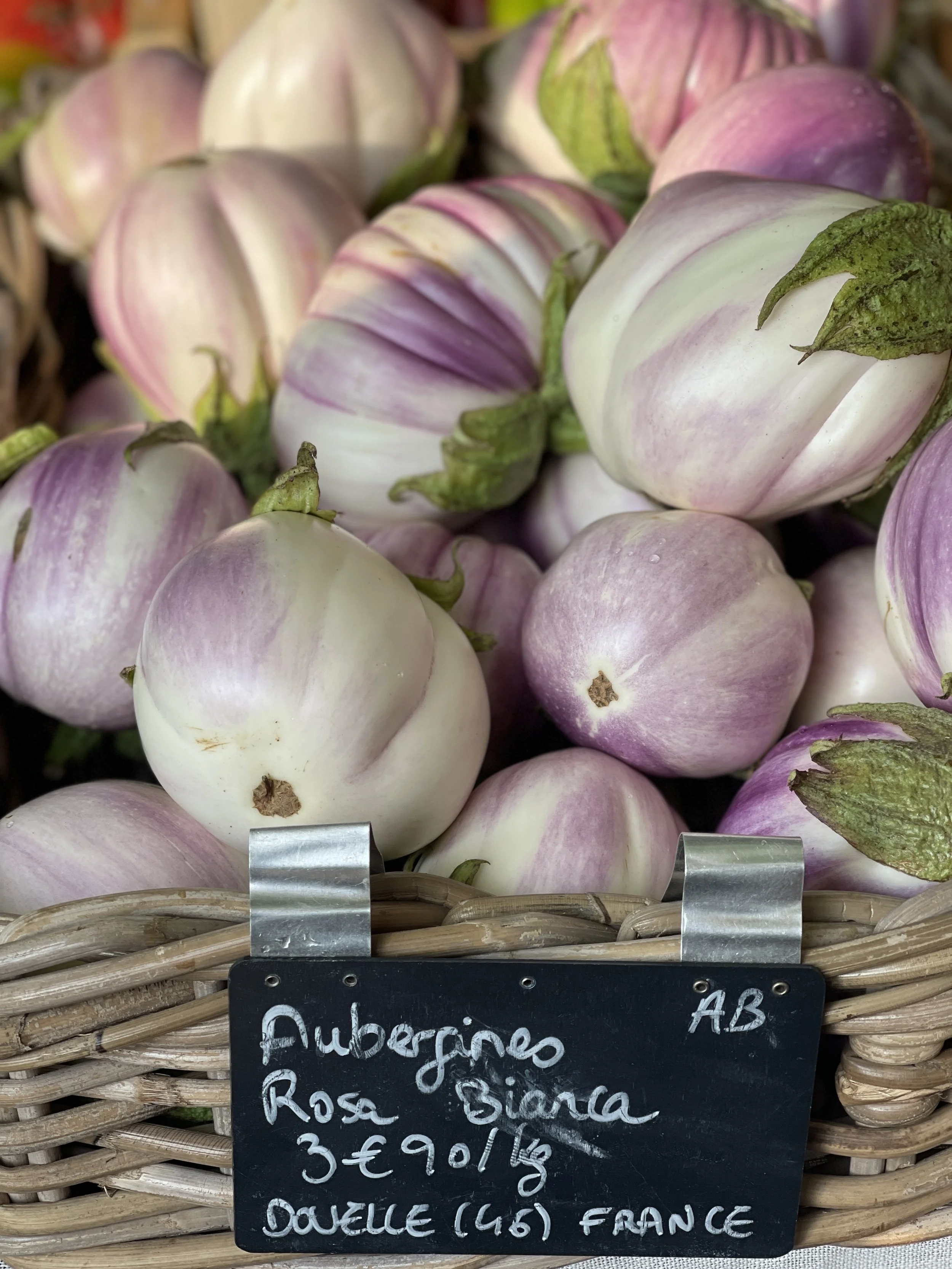 A basket of white and purple-striped eggplants with a handwritten sign in front that reads: 'Aubergine Rosa Bianca 3€ 90/1 kg DOUCE (46) FRANCE'.