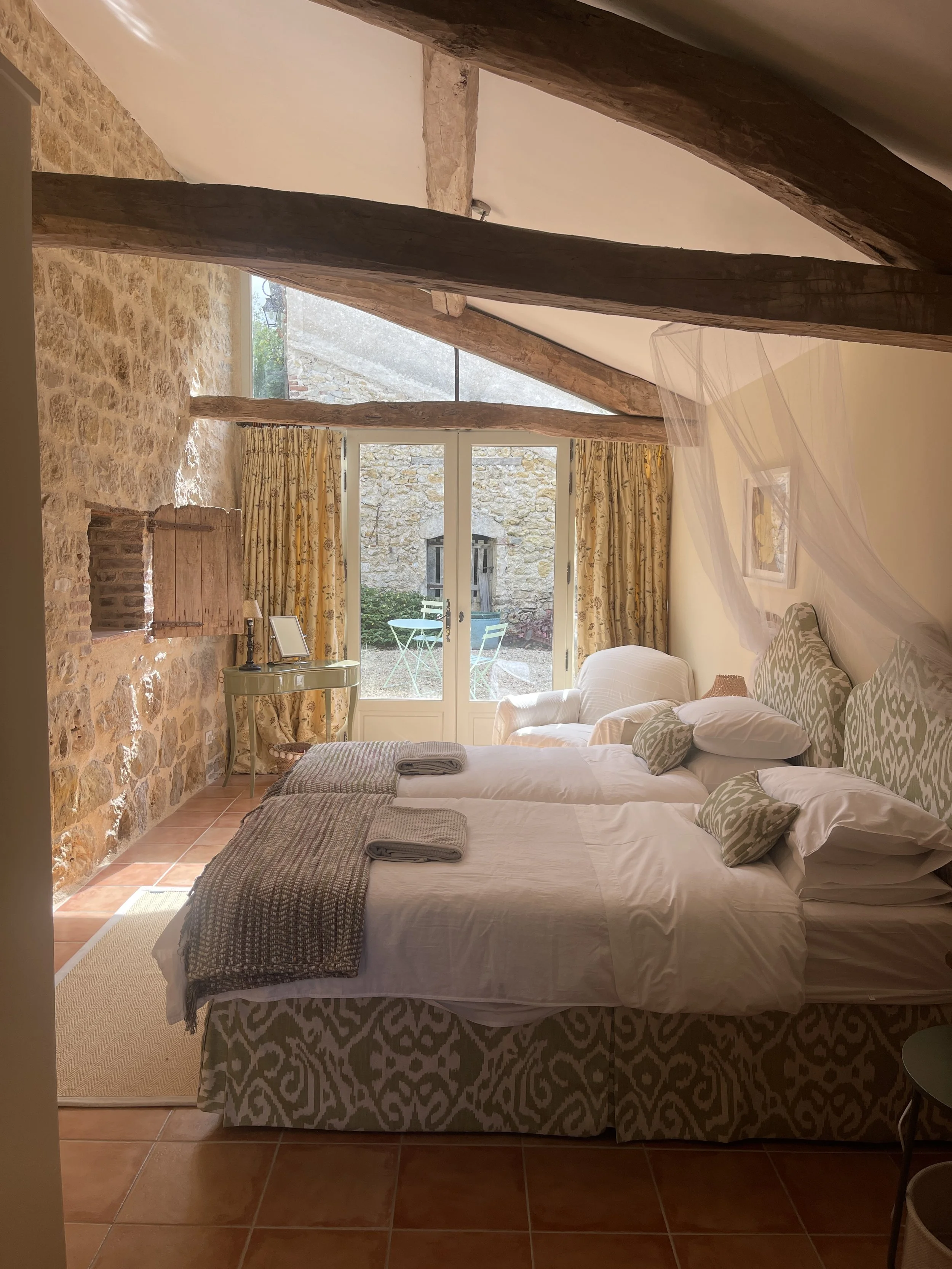 A cozy bedroom featuring two twin beds with patterned headboards, a window with yellow floral curtains, stone and wooden wall accents, and a view of a small outdoor patio with a table and chairs.