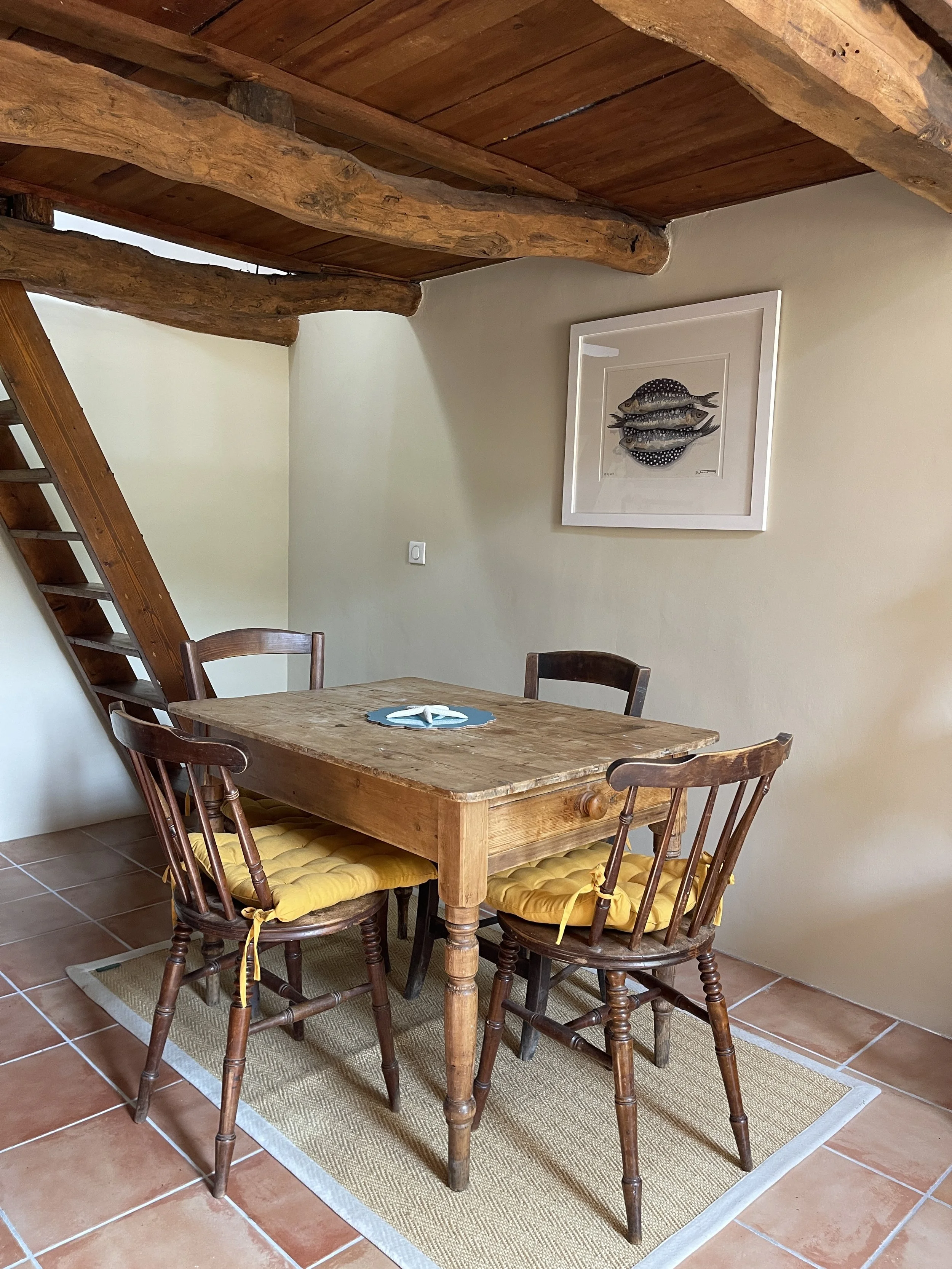 A rustic dining area with four wooden chairs with yellow cushions around a wooden table, on a beige rug, with a wall art of fish above, and a staircase leading up on the left, and a wooden ceiling with exposed beams.