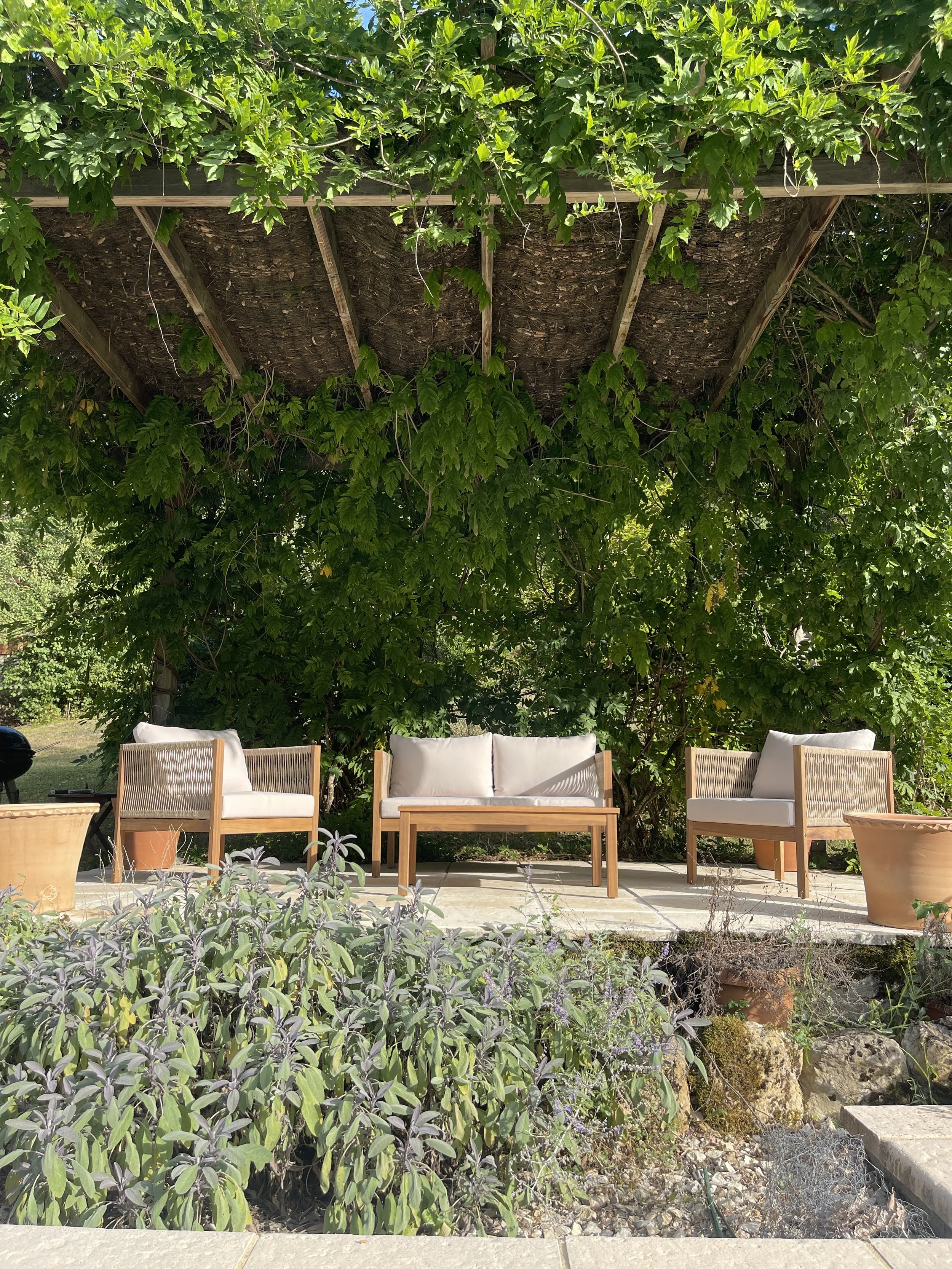 A patio area with outdoor wicker furniture including a loveseat and two single chairs, with cushions on each piece. The furniture is arranged in front of a lush green hedge and underneath a wooden pergola covered with vine leaves, creating a shaded s