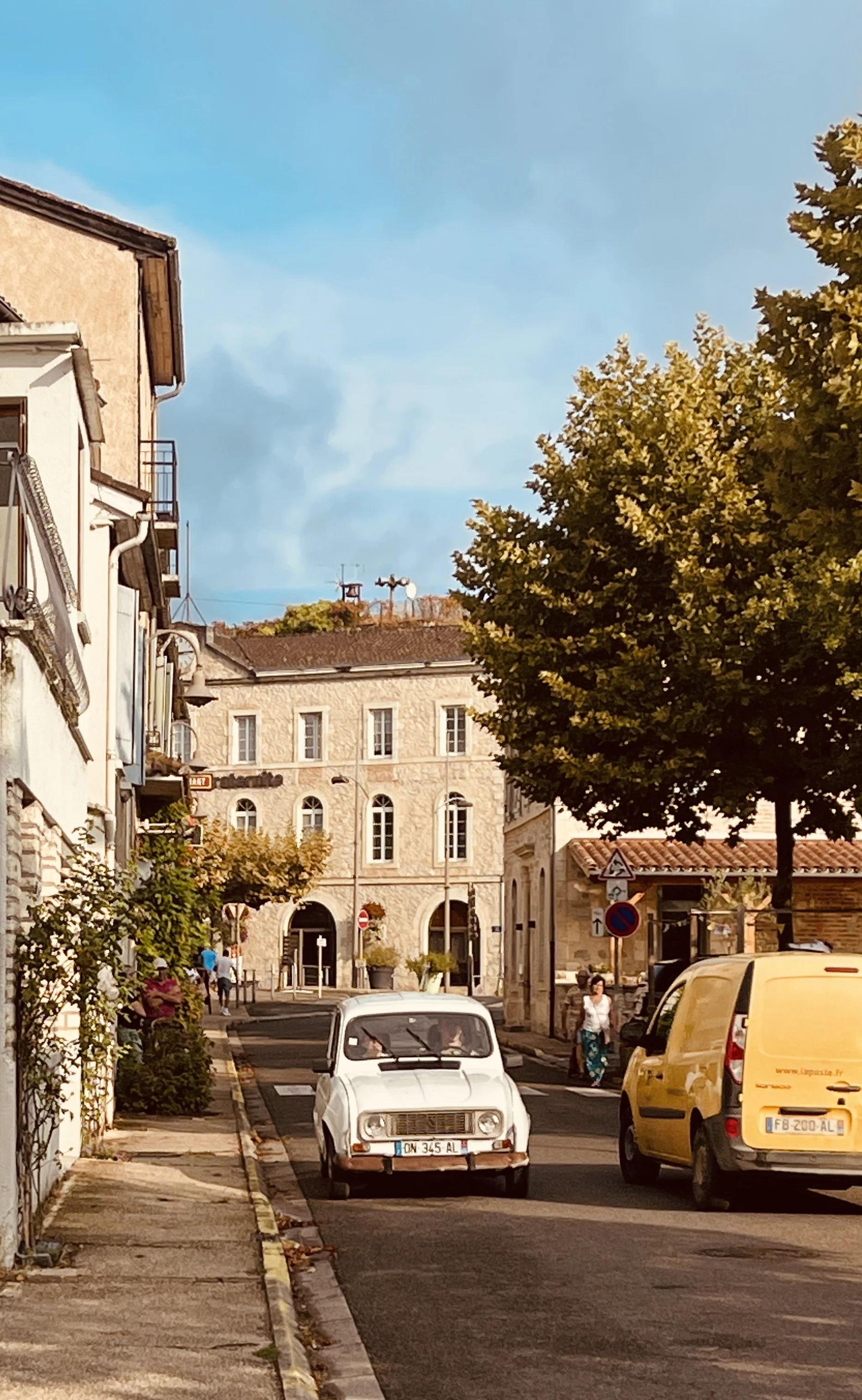Street scene with parked cars, people walking, trees, and historic buildings, captured under a blue sky.