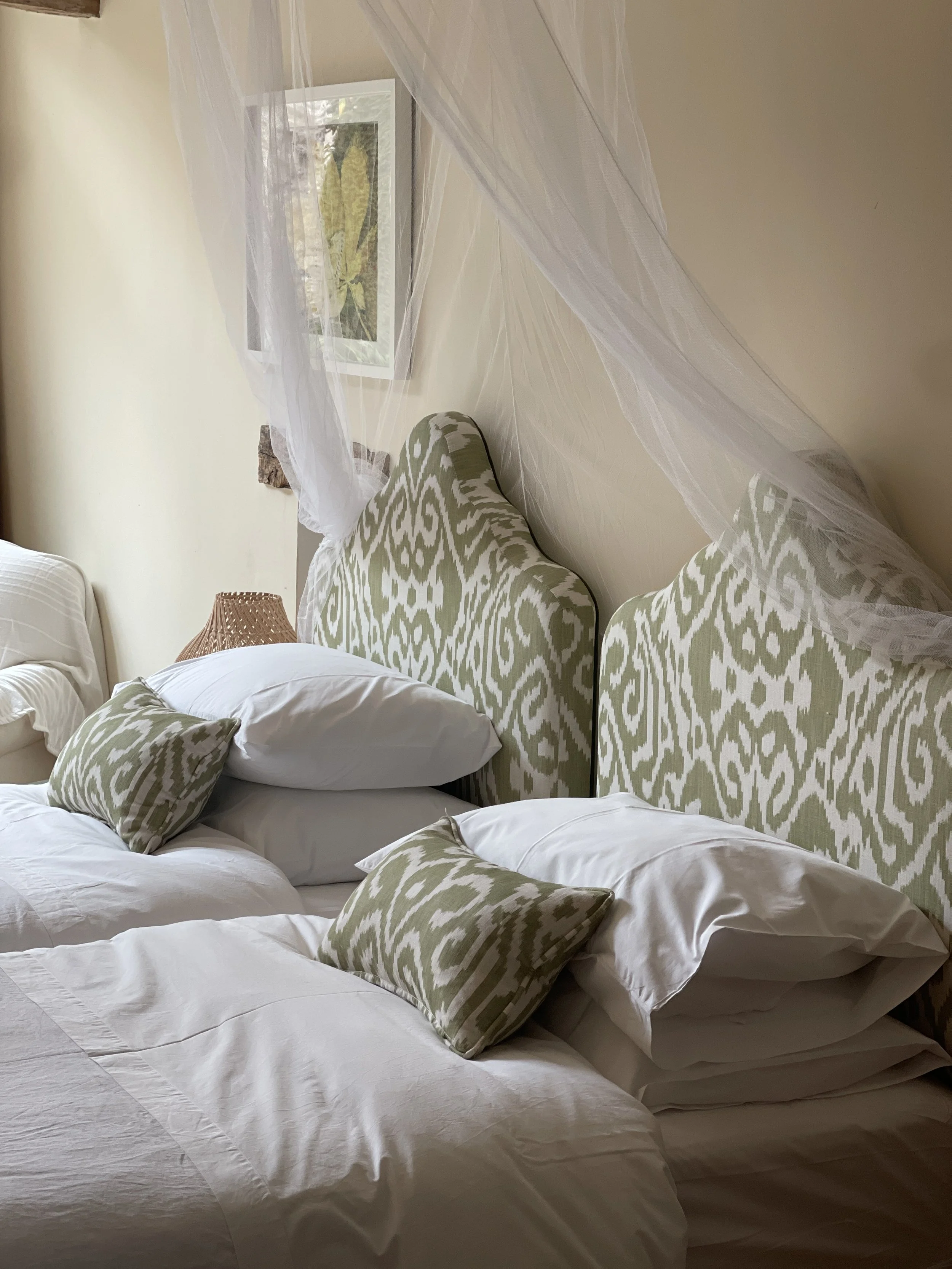 A bedroom with two beds pushed together, two beds with white sheets and pillows, decorated with green and white patterned headboards and matching pillows, a framed botanical picture on the wall, and a sheer white canopy draped around the headboards.