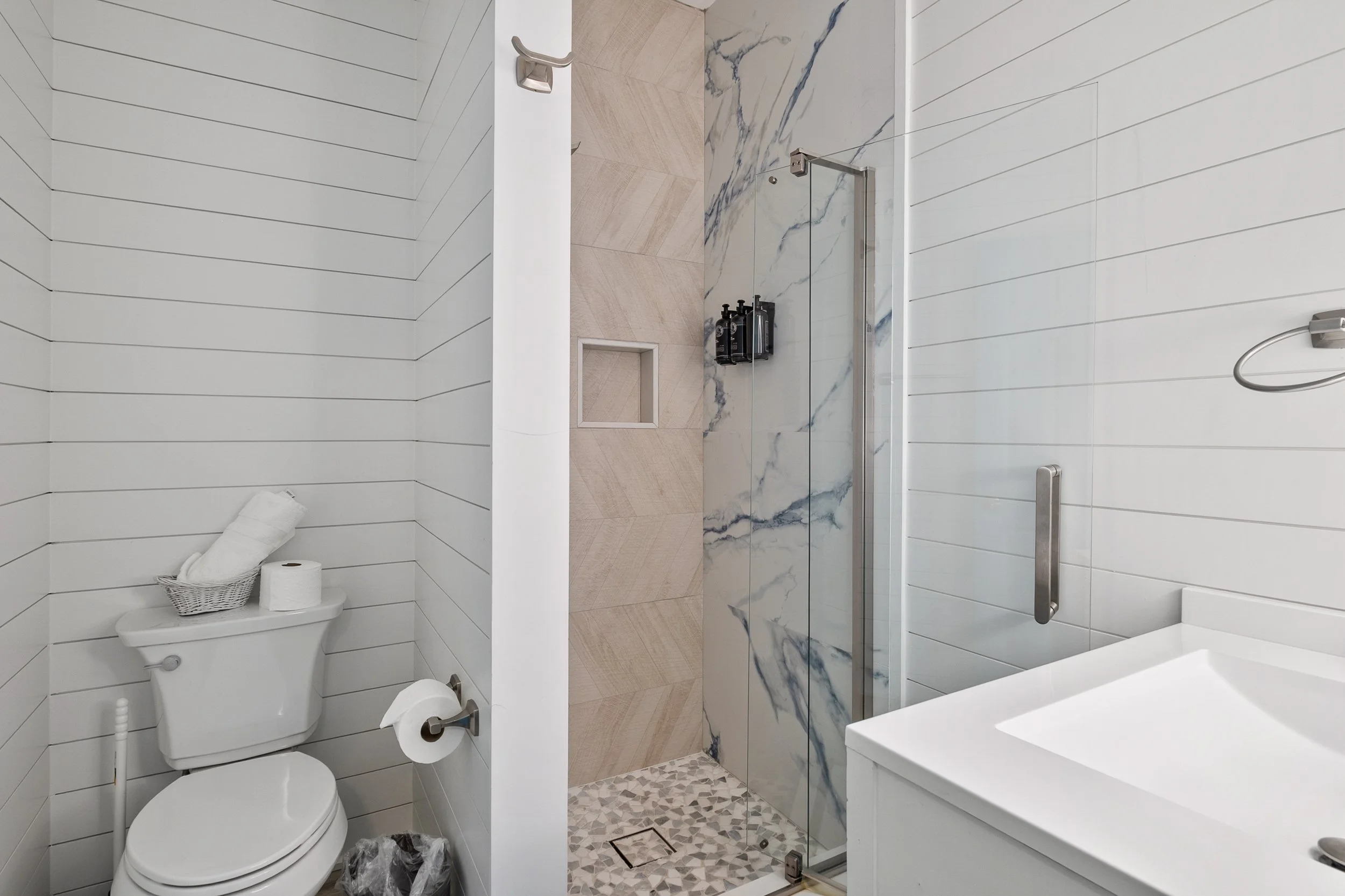 Modern bathroom with white horizontal shiplap walls, a toilet with a basket and toilet paper on top, a walk-in shower with marble and beige tile walls, and a pebble-tile floor.