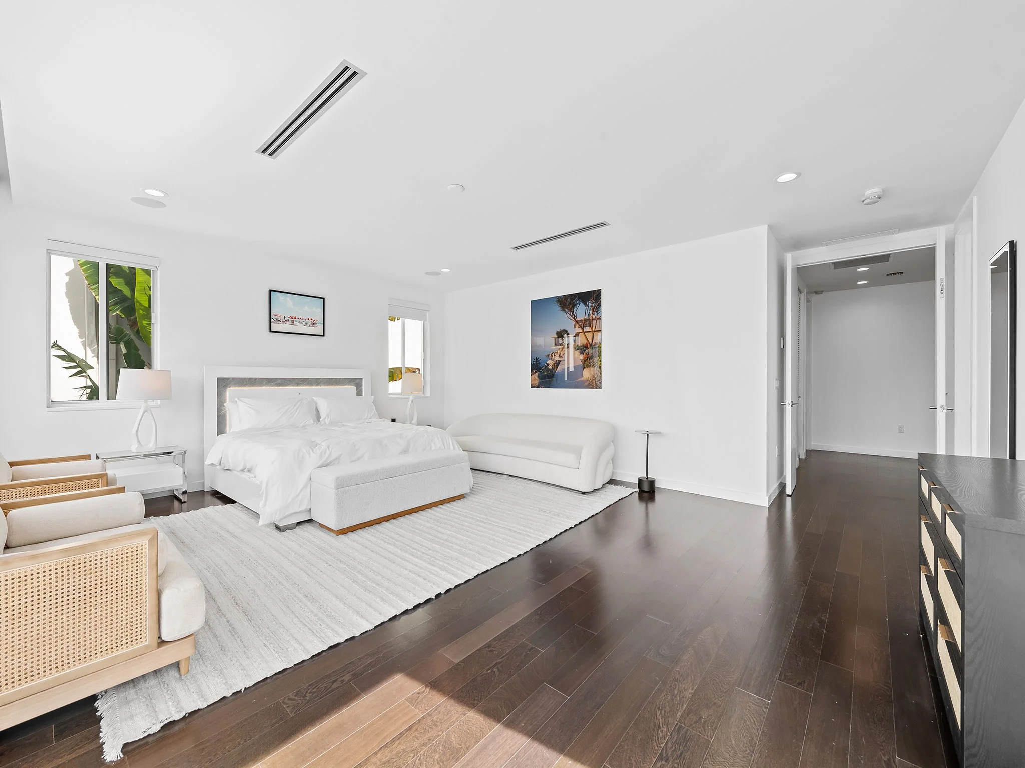 Bright white bedroom with large bed, framed pictures on wall, wooden furniture, and dark wooden floor.