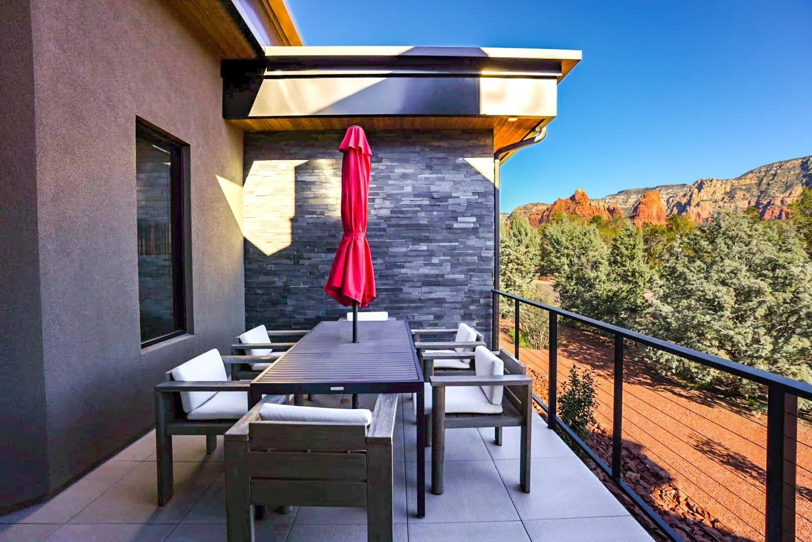 Balcony with outdoor dining table, six white chairs, closed red umbrella, gray stone accent wall, forested landscape, red rock formations in the distance, clear blue sky.