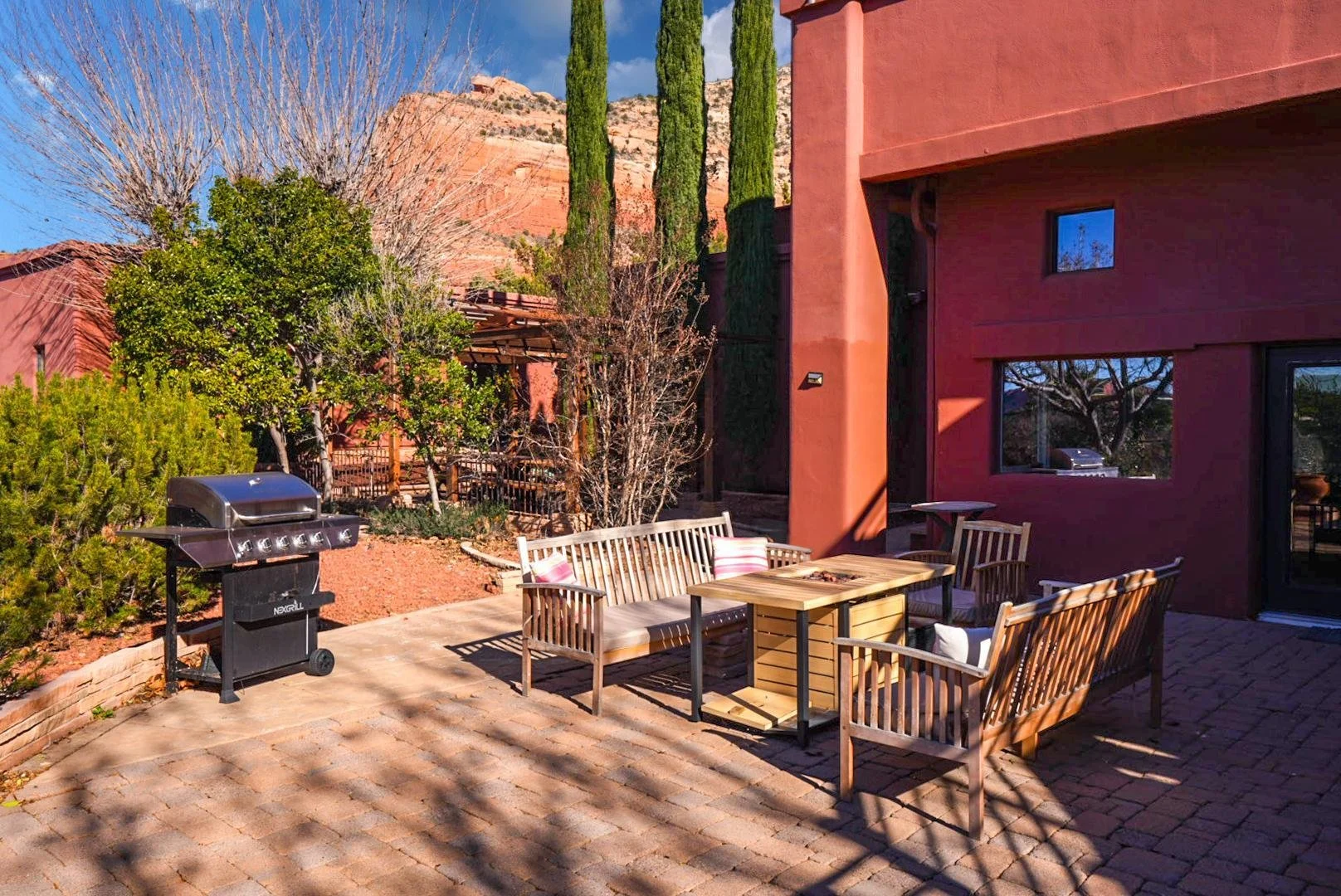 Outdoor patio area with patio furniture, a grill, and surrounding trees, in front of a red building with cypress trees and rocky hills in the background.