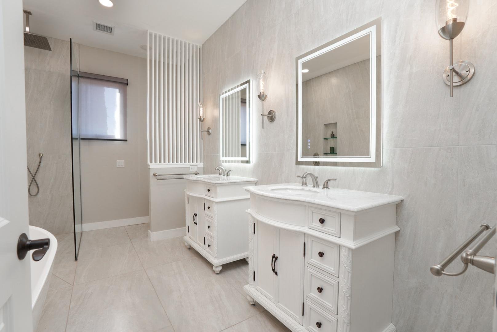 Modern bathroom with two white vanities, illuminated mirrors, and a walk-in shower with glass door.