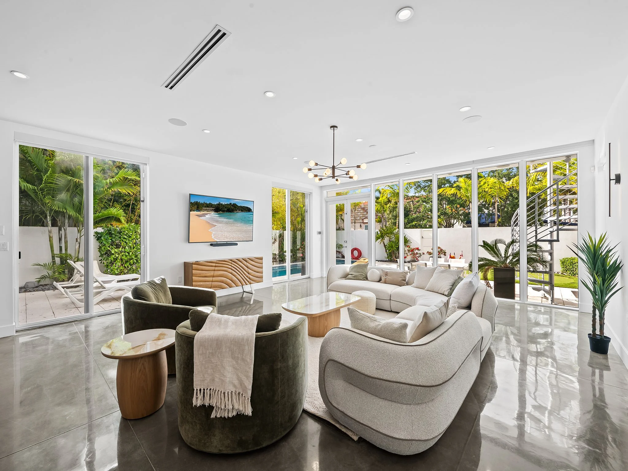 Bright living room with large glass doors and windows open to a lush outdoor garden. Modern furniture includes a curved white sofa, gray armchairs, and small wooden side tables. A flat-screen TV is mounted on the wall, and a contemporary chandelier h