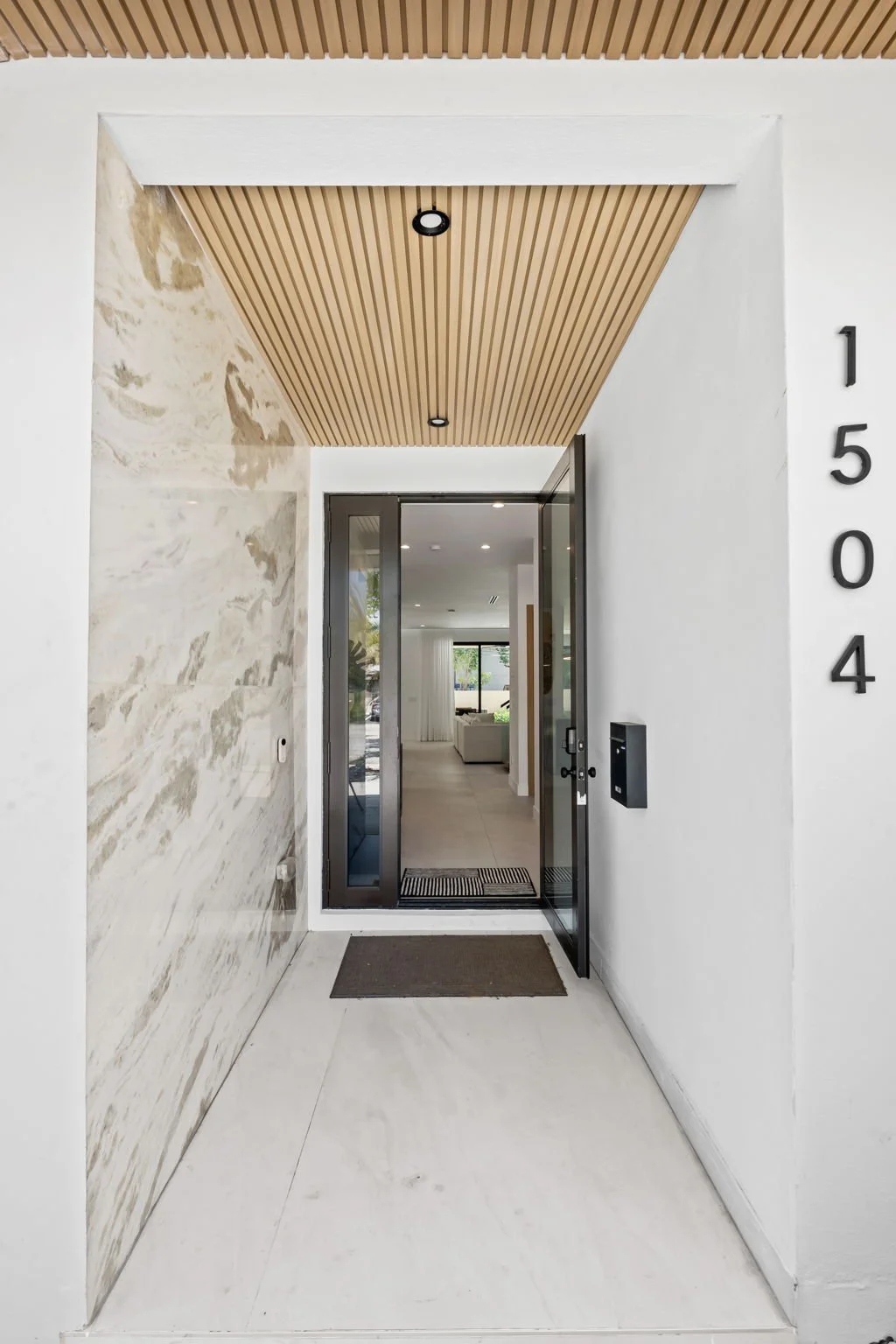 Modern building entrance with white walls, a marble accent wall, a wooden slat ceiling, a glass-paneled door, and house number 1504 displayed vertically on the right side.