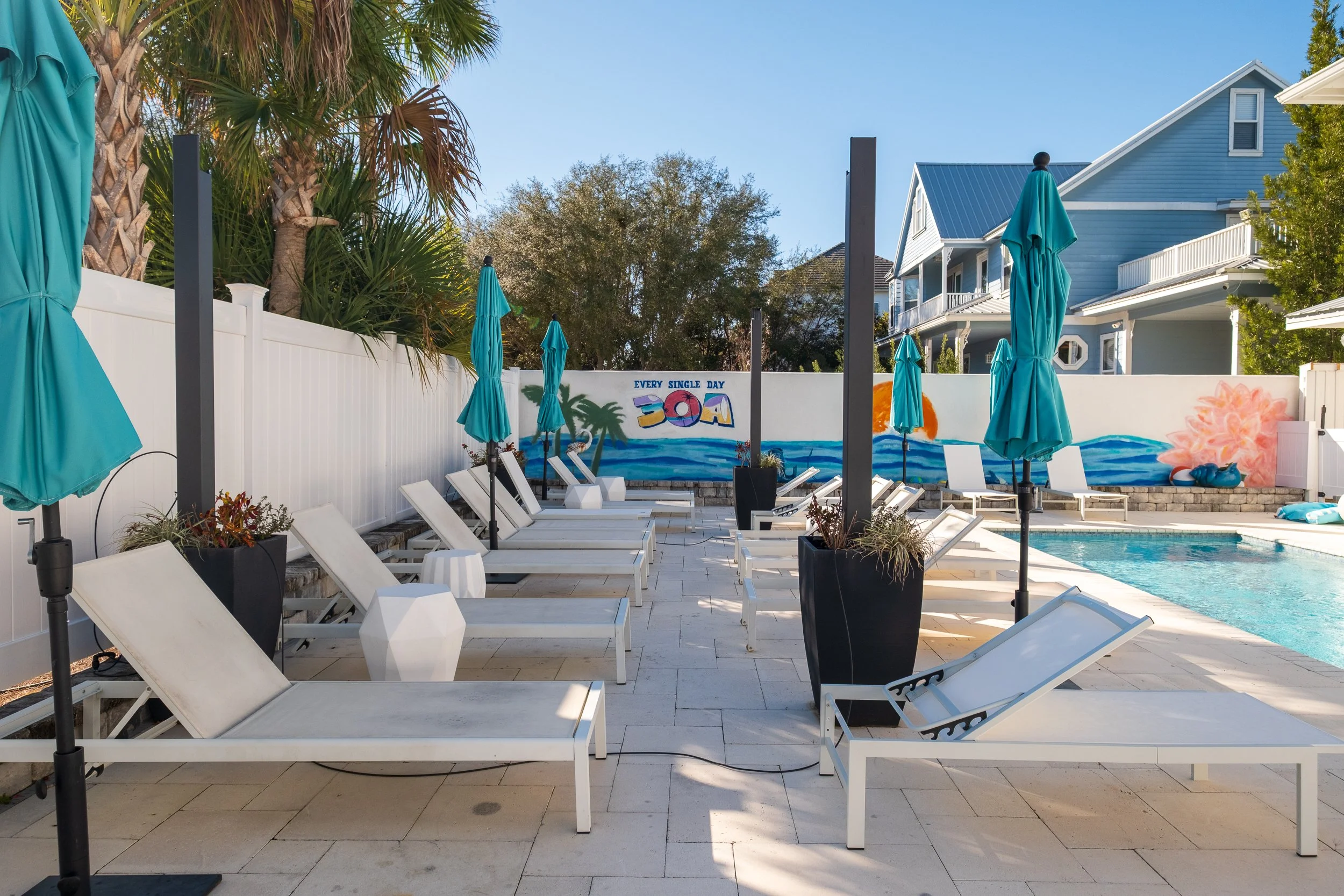 Empty swimming pool area with white lounge chairs, blue umbrellas, and a colorful mural on a white fence with palm trees and a house in the background.
