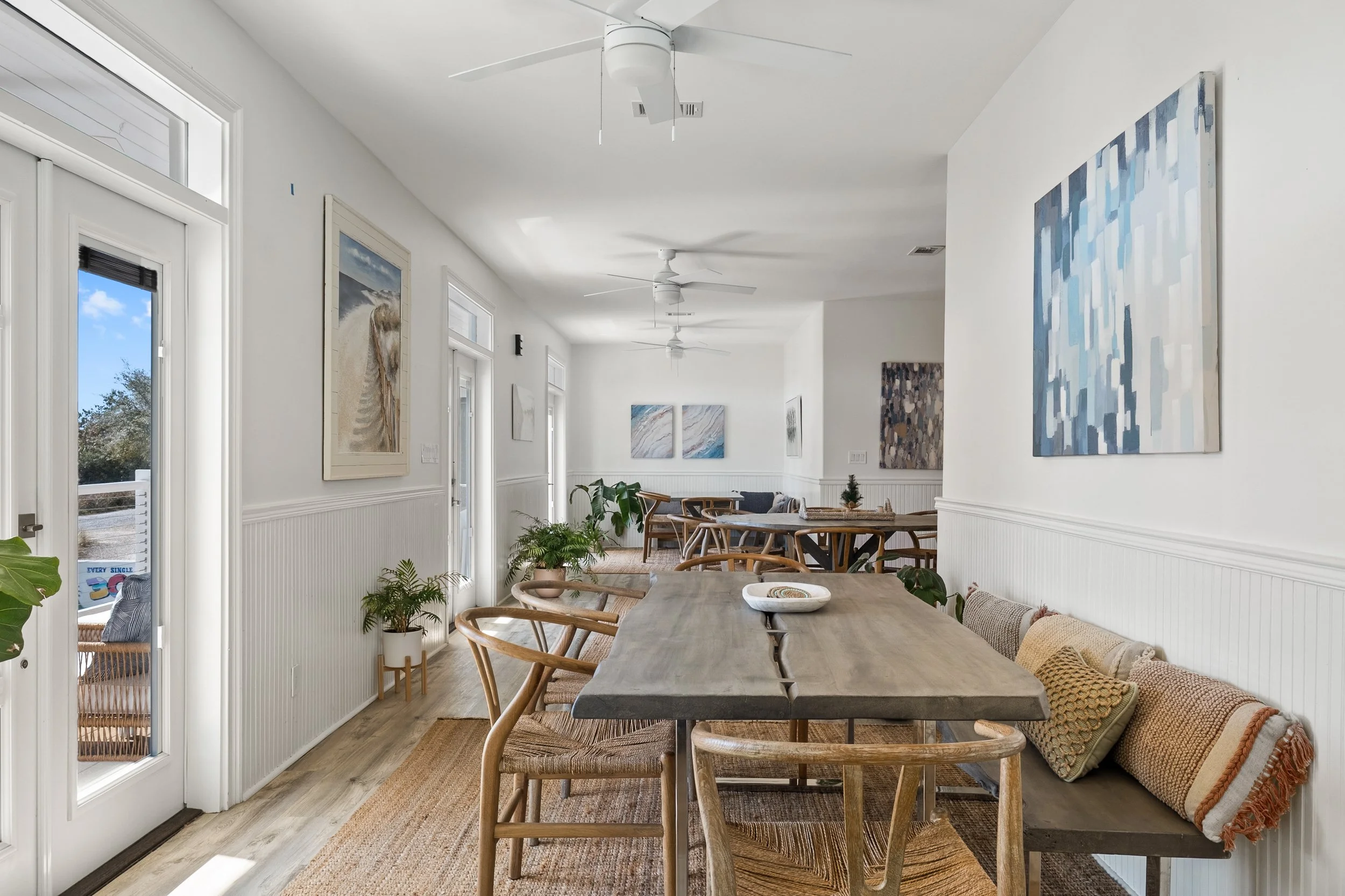 Open dining area with a wooden table, chairs, wall art, and potted plants, featuring white walls, natural light, and ceiling fans.