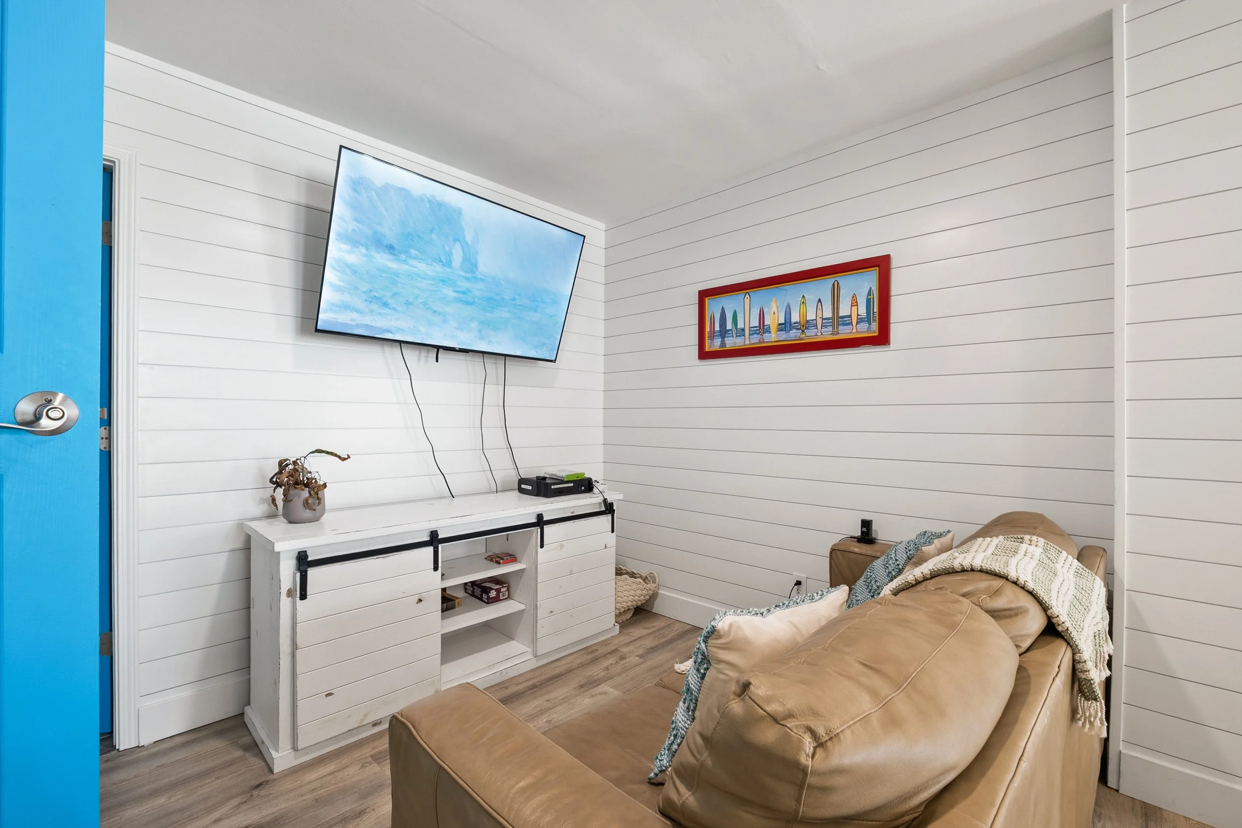 Living room with white wood-paneled walls, a tan leather sofa, a white TV stand, a wall-mounted flat-screen TV, and a colorful framed art piece featuring surfboards, with hardwood flooring.