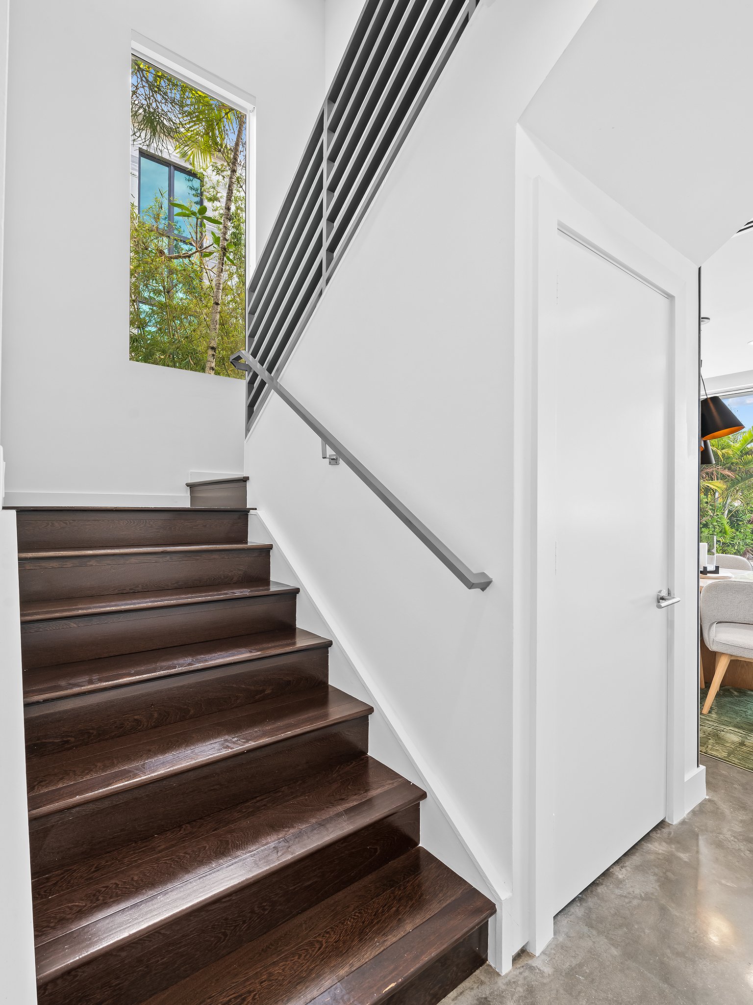 Interior view of a staircase with dark wooden steps and a white wall. A window showing green trees and blue sky is visible at the top of the stairs. There is a metal railing along the stairs.