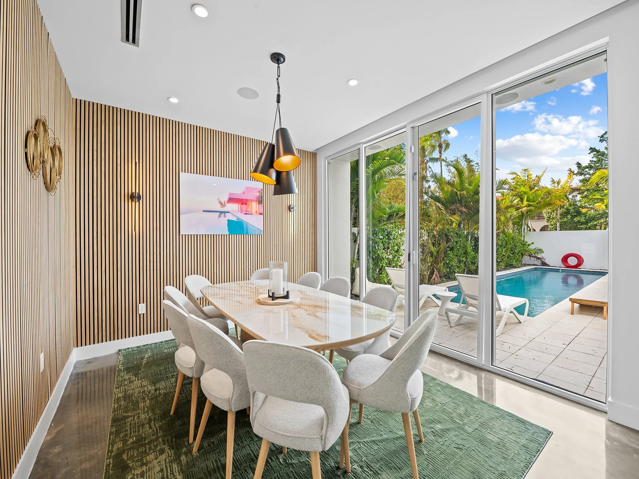 Modern dining room with beige upholstered chairs around a marble table, green rug, wooden accent wall, large glass sliding doors opening to a pool and lush greenery outside, pendant lights, and a wall-mounted TV.