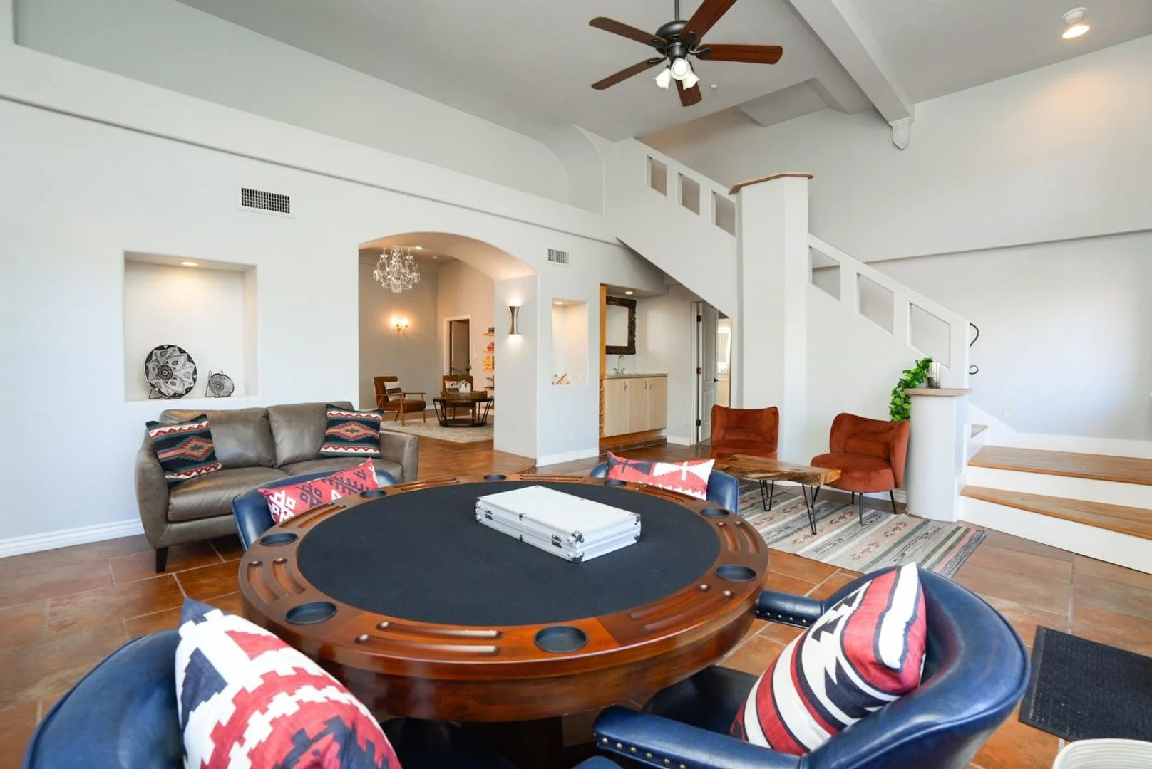 Living room with a poker table, a leather sofa with patterned pillows, two red chairs, a wooden staircase, and a balcony view. Interior decorated in a modern style with a white ceiling, ceiling fan, and accents.