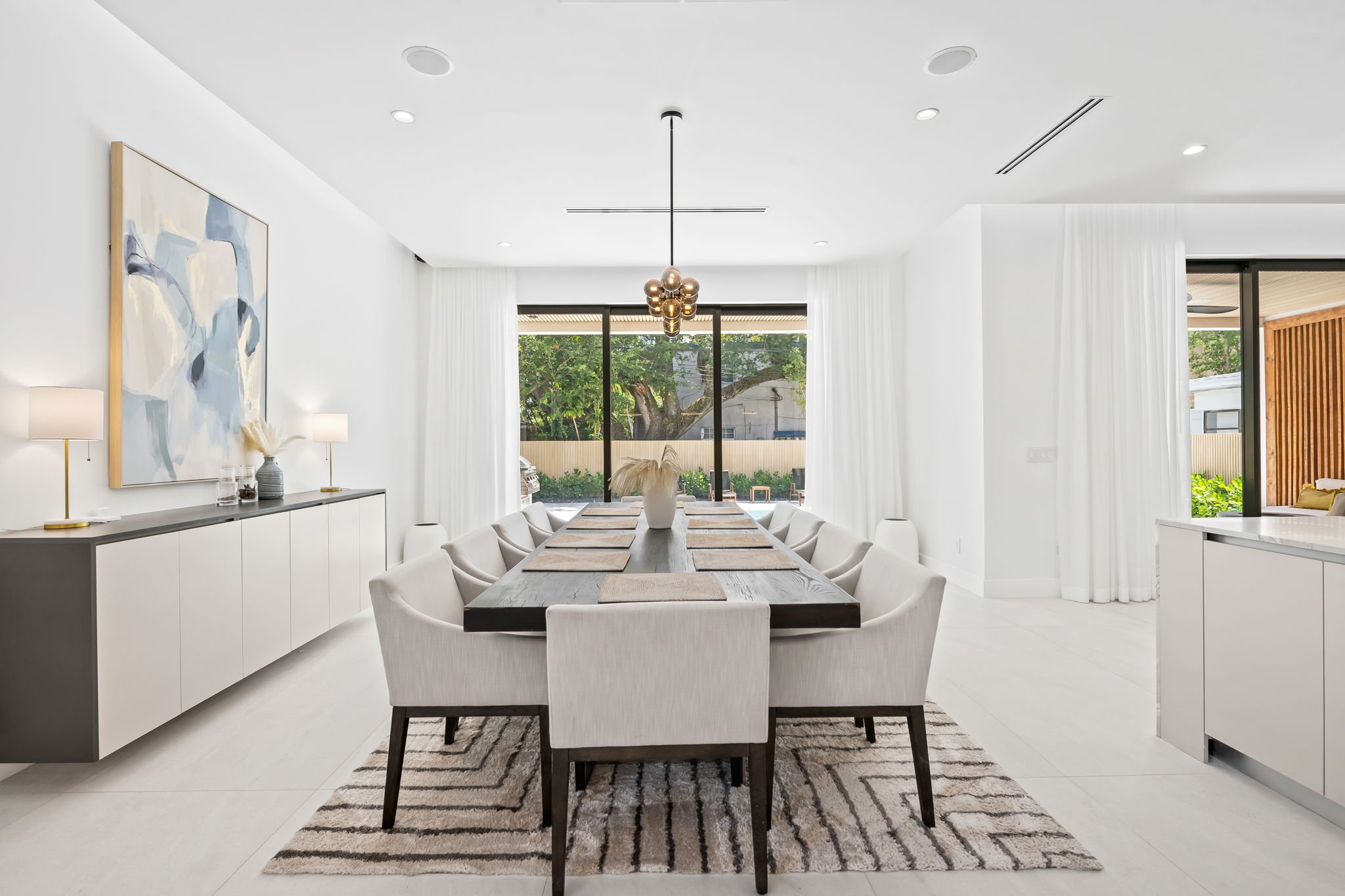 Sunlit dining room with long wooden table, upholstered chairs, and contemporary artwork — Sun Haven Collection luxury vacation home.