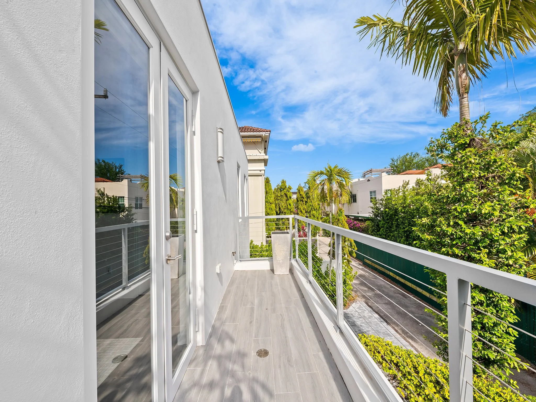 Balcony with glass sliding door, white exterior walls, gray tiled floor, and a view of trees, bushes, and a street with cars below, under a partly cloudy blue sky.
