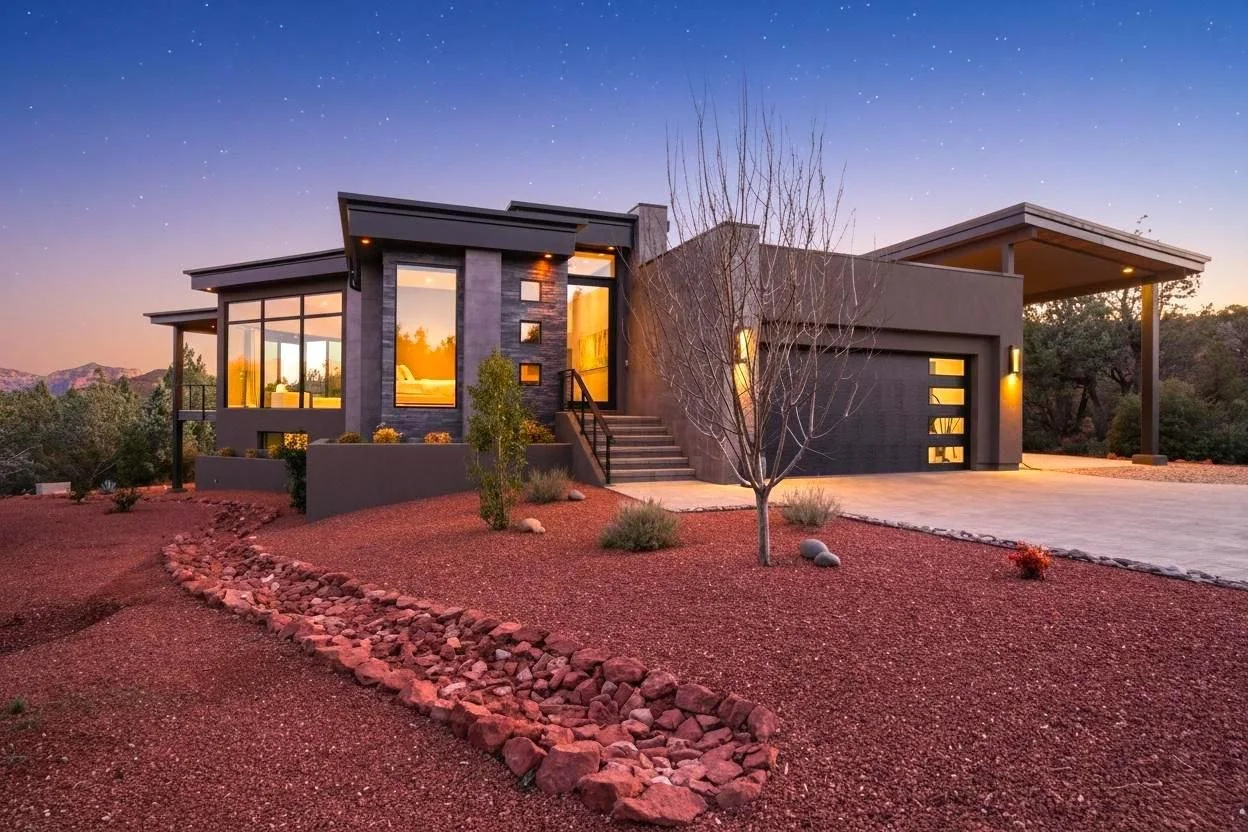 Modern house with large windows, set in a desert landscape at sunset, surrounded by rocks, sparse trees, and desert plants, with a paved driveway.