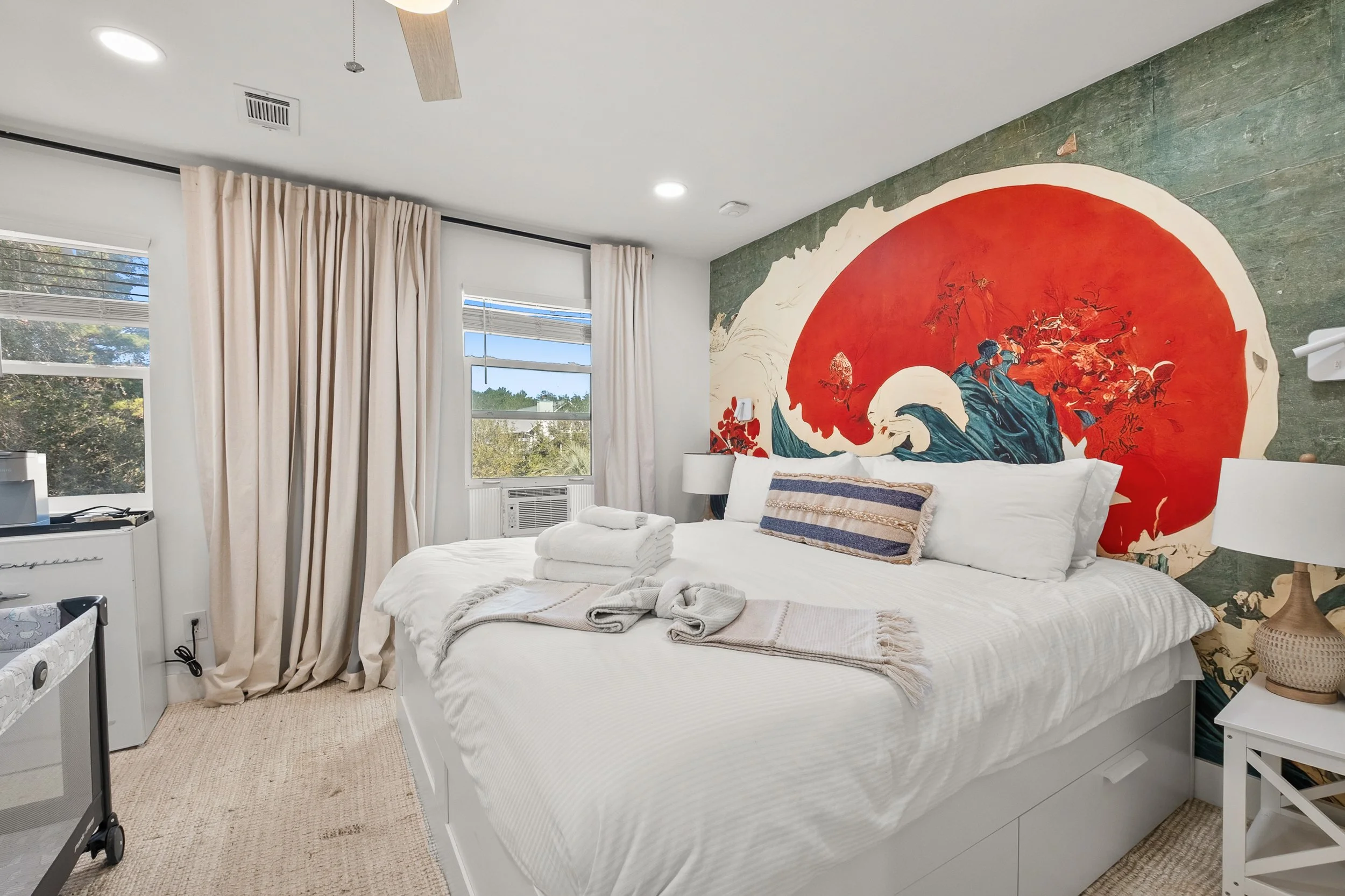 Cozy bedroom with a large bed, white bedding, and decorative pillows, featuring a Japanese wave mural on the headboard wall, surrounded by beige curtains and a window with a view of trees.