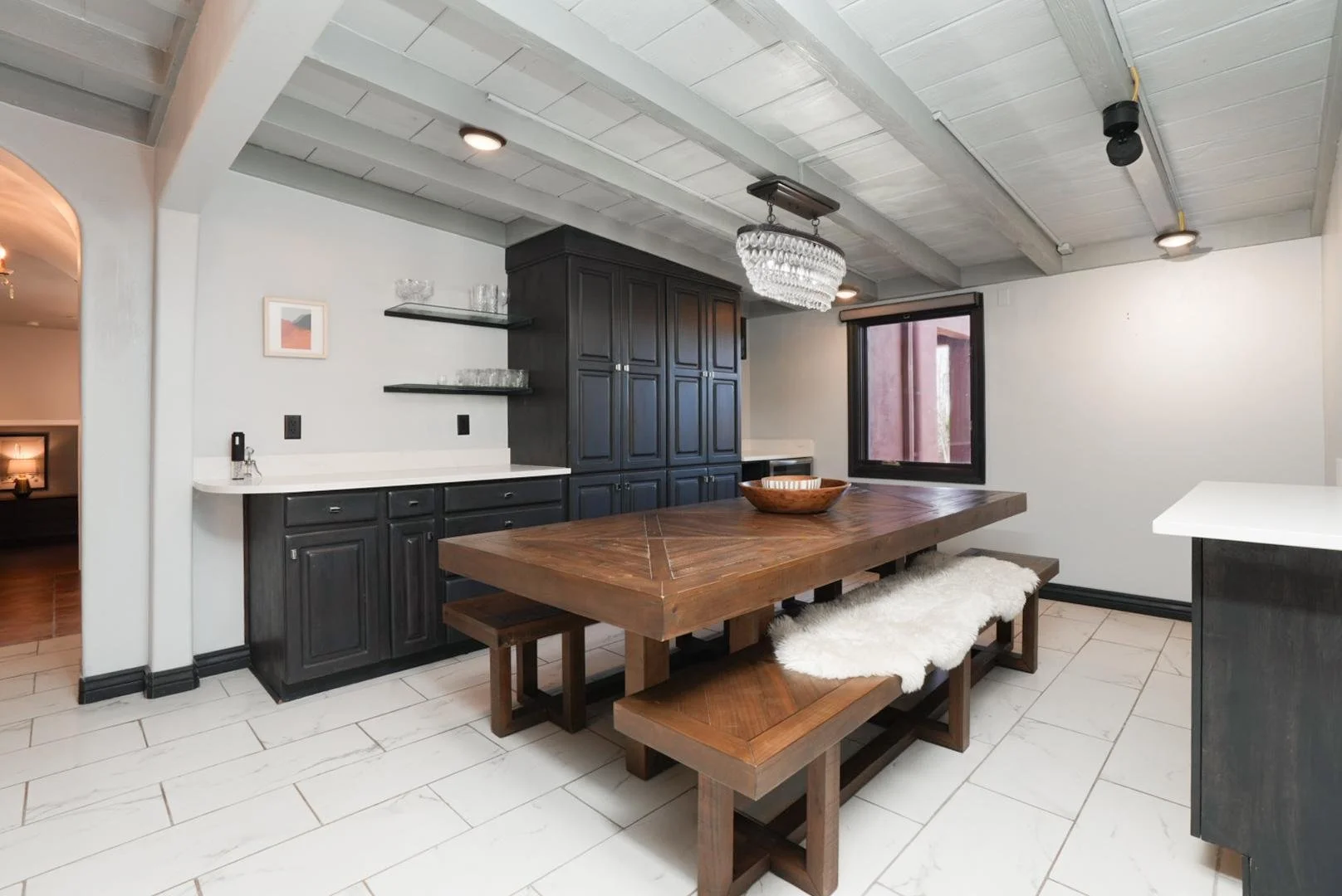 Dining room with a large wooden table, black cabinets, shelf with glassware, a chandelier, window, and a bench with a white fur cushion.