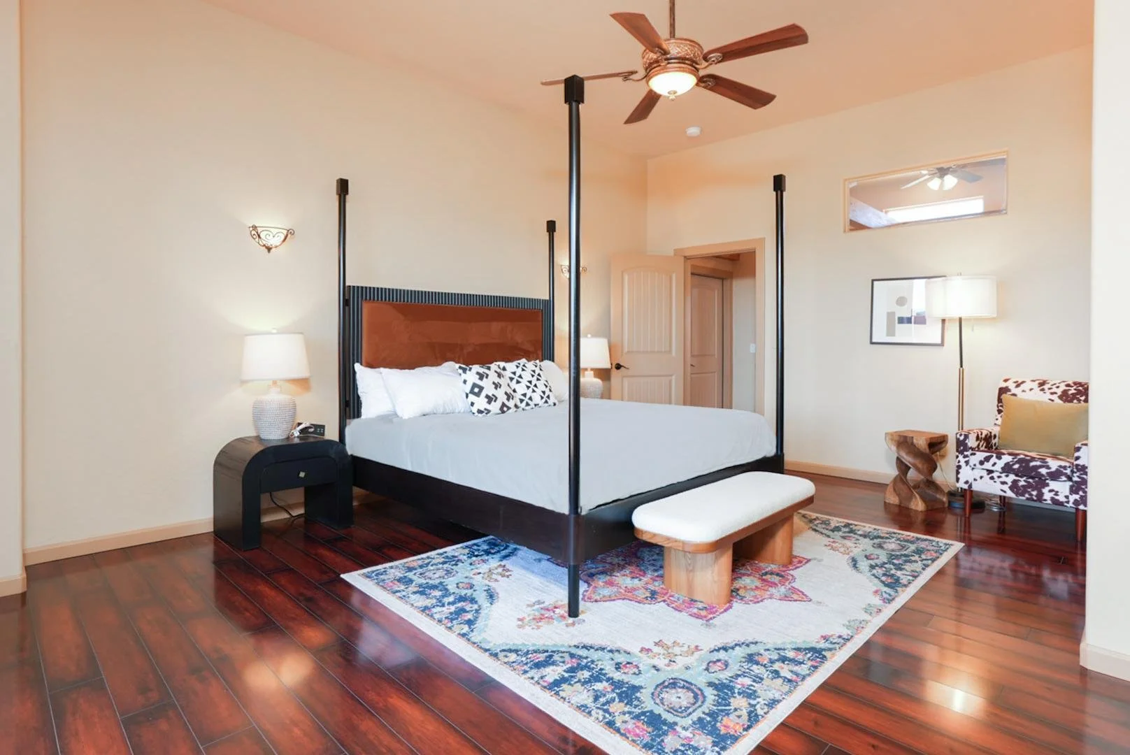 A bedroom with a four-poster bed, wooden flooring, a patterned area rug, two bedside lamps, a cowprint armchair, and a bench at the foot of the bed.