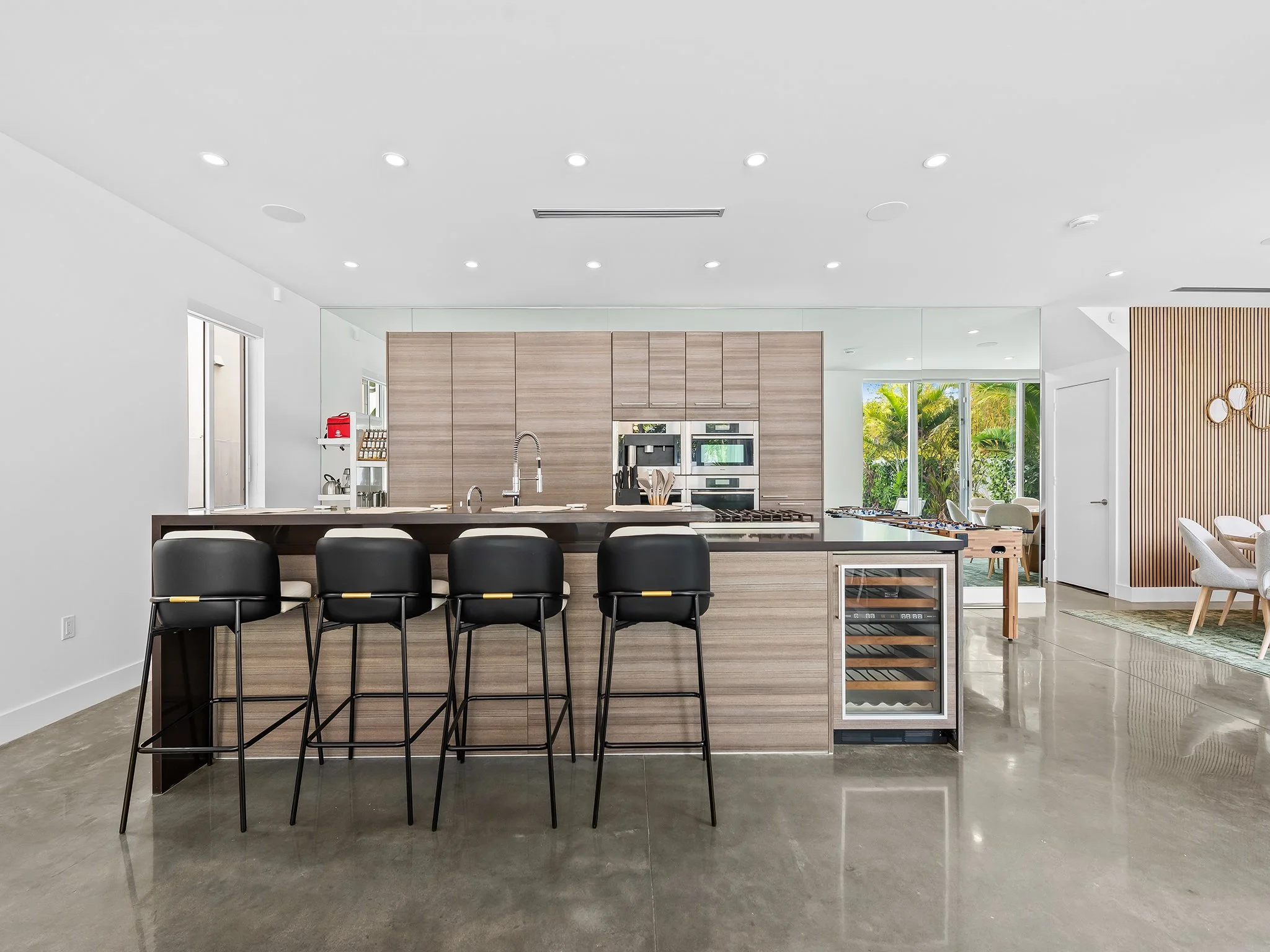Modern kitchen with light wood cabinets, black bar stools at a kitchen island, stainless steel appliances, and large windows overlooking greenery. Bright white walls and ceiling with recessed lighting.