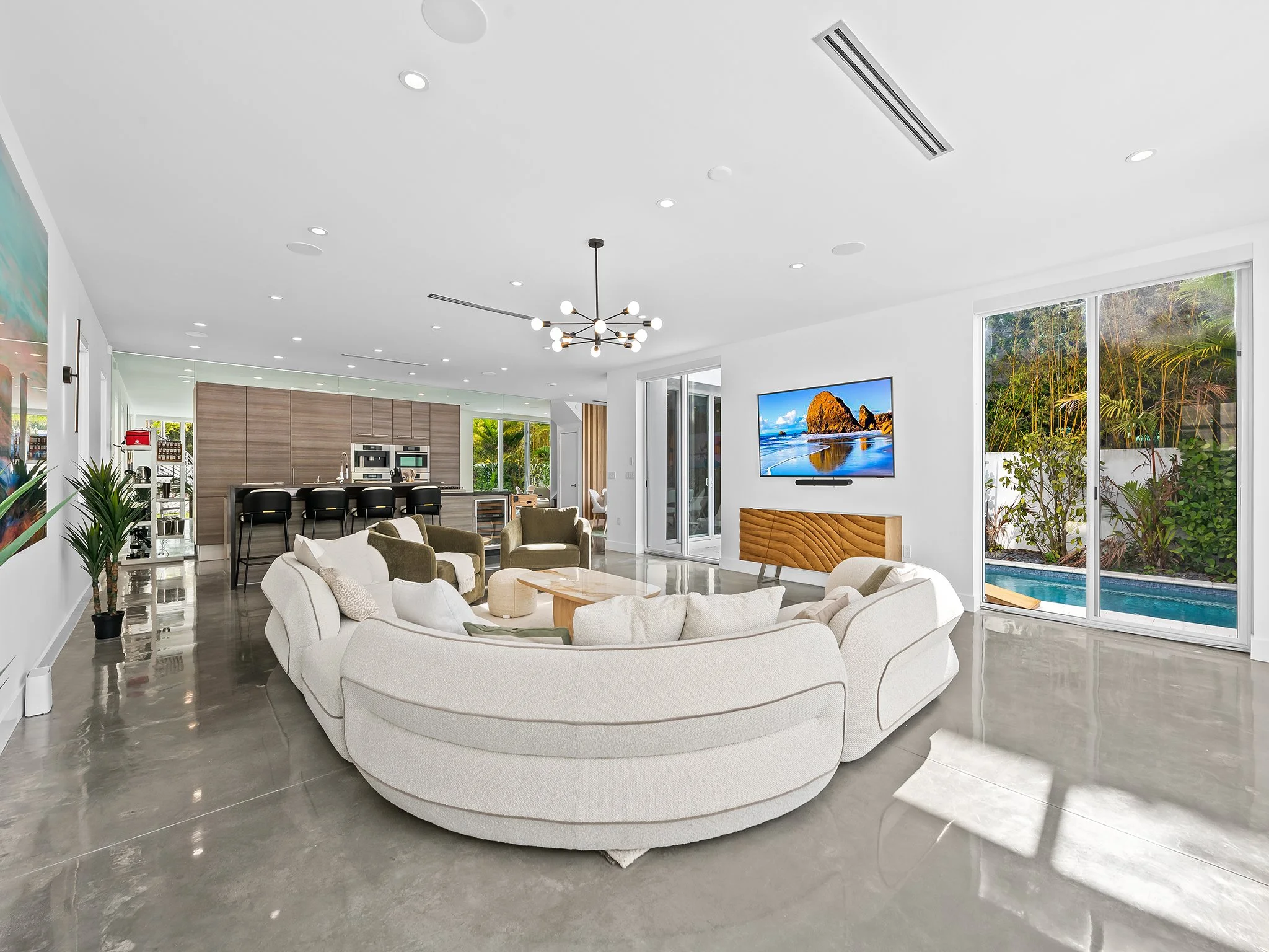 Modern open plan living room with white circular sofa, TV mounted on a white wall, large sliding glass doors with view of outdoor plants and pool, and contemporary ceiling lights.