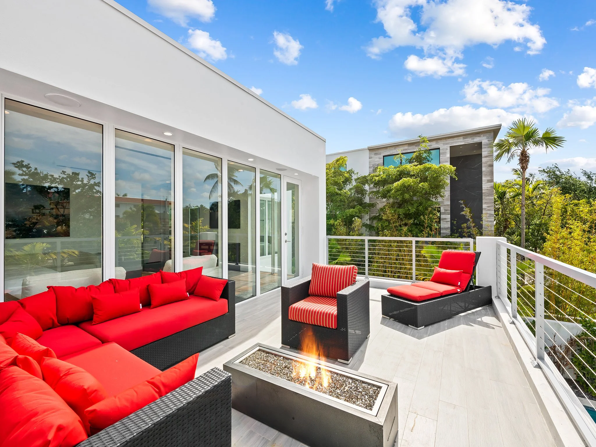 A modern outdoor terrace with red cushioned seating, a fire pit, and glass doors leading inside, surrounded by lush greenery under a partly cloudy blue sky.