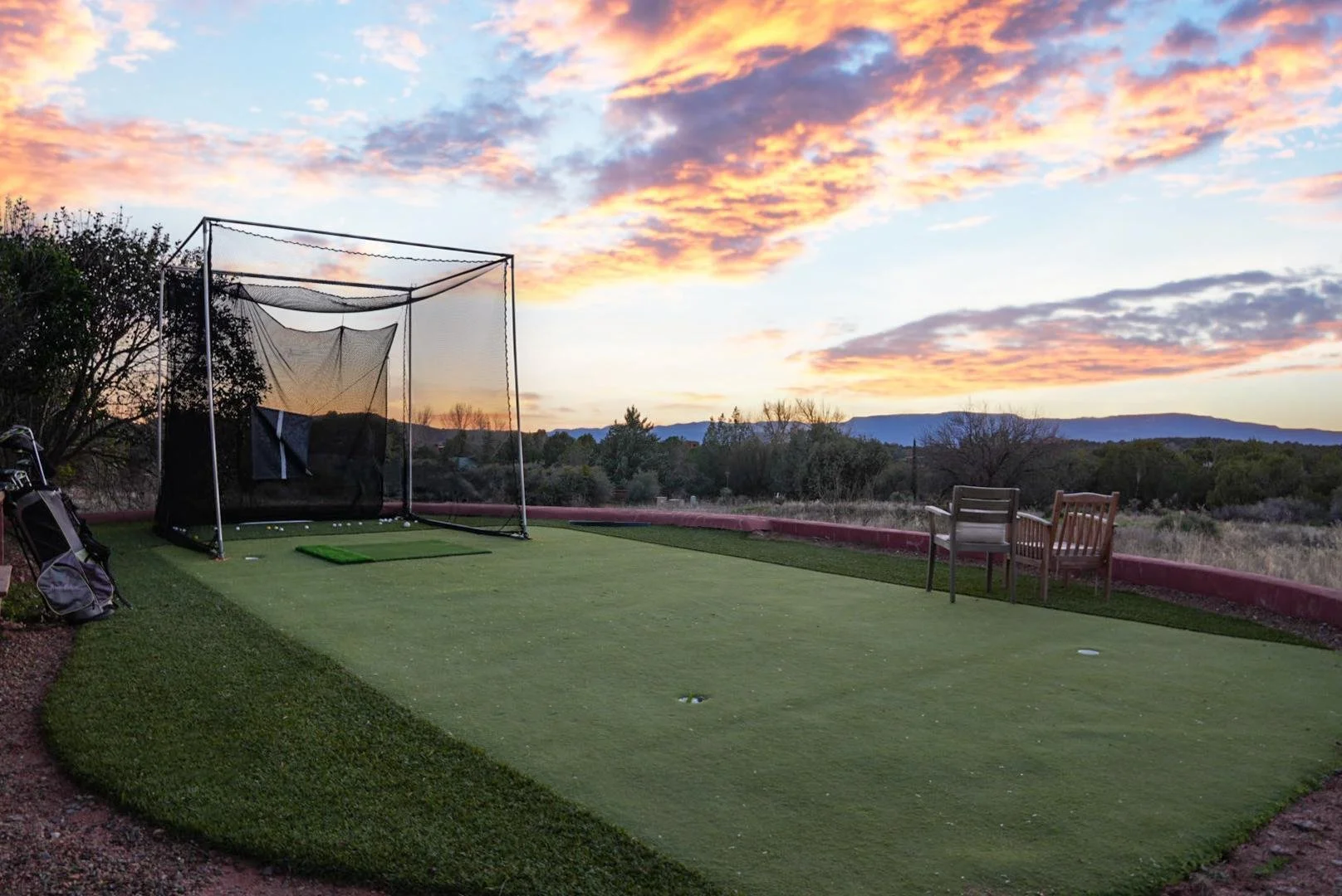 A golf practice area with a putting green, surrounded by a low border, outdoor chairs, a golf bag, and a golf net, with a colorful sunset sky and mountains in the background.