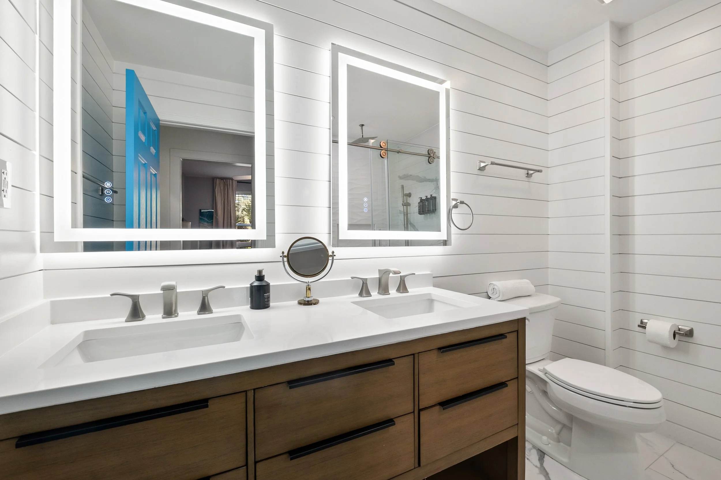 Modern bathroom with white paneled walls, double vanity with white countertop, two illuminated mirrors, and a toilet. There is a glass shower enclosure visible in the mirror reflection.