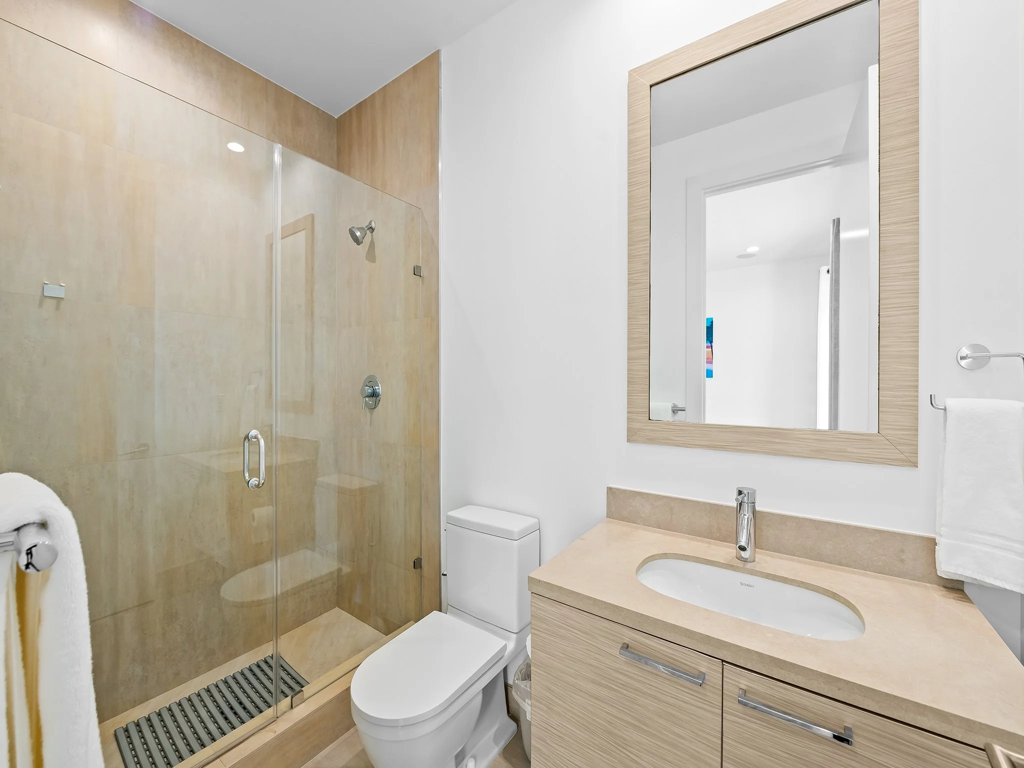 A modern bathroom with a glass shower enclosure, a toilet, and a wooden vanity with a mirror. The shower has beige tiles and a ceiling-mounted showerhead. There is a white towel hanging on a towel holder.
